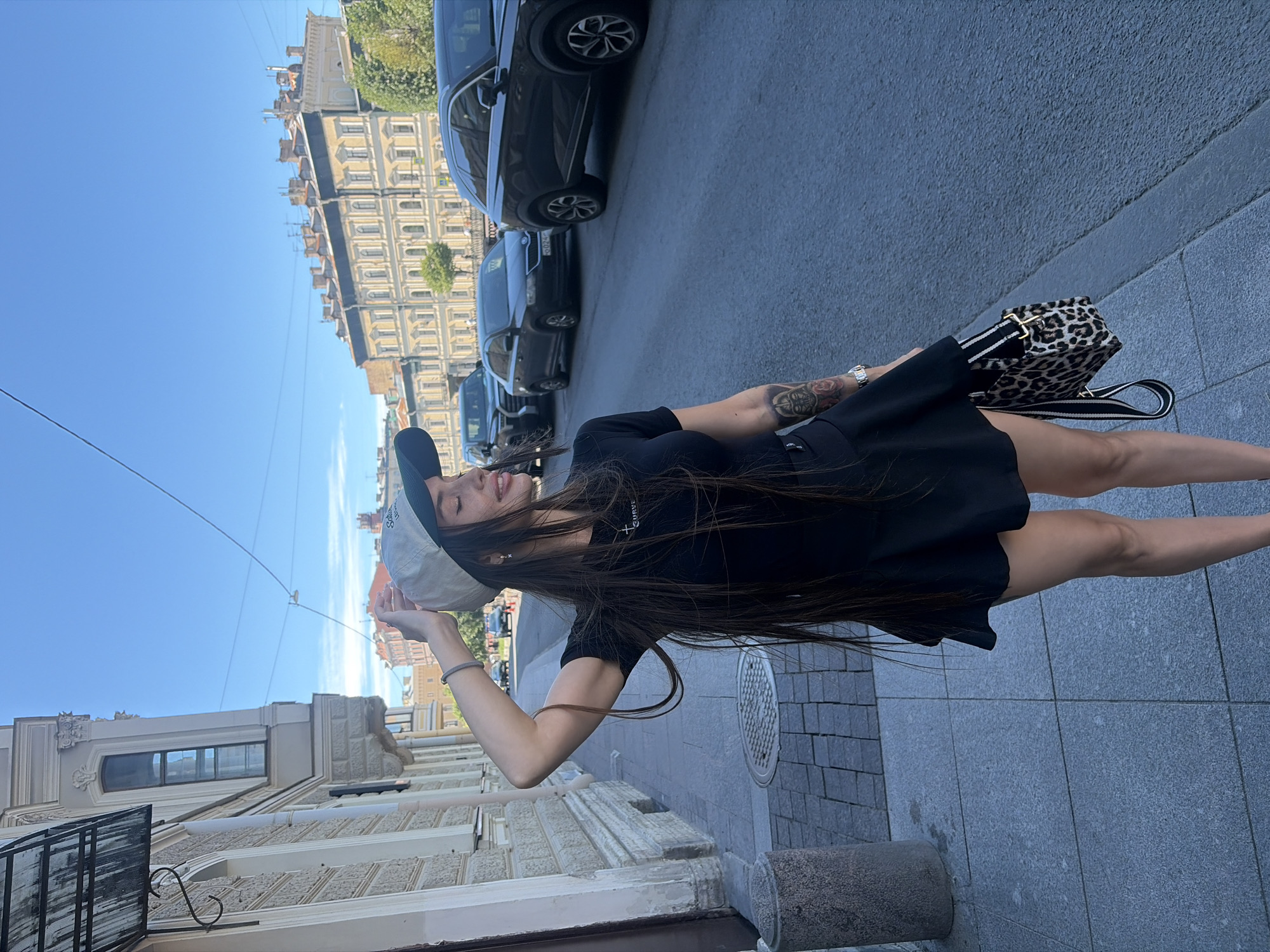 A young woman stands on a sidewalk in Saint Petersburg, Russia, looking to her right with her tongue sticking out slightly. She is wearing a black t-shirt dress, a baseball cap, and a leopard-print purse. The street is lined with cars, and classic St. Petersburg architecture is visible in the background. The sky is clear and blue, indicating a sunny day.