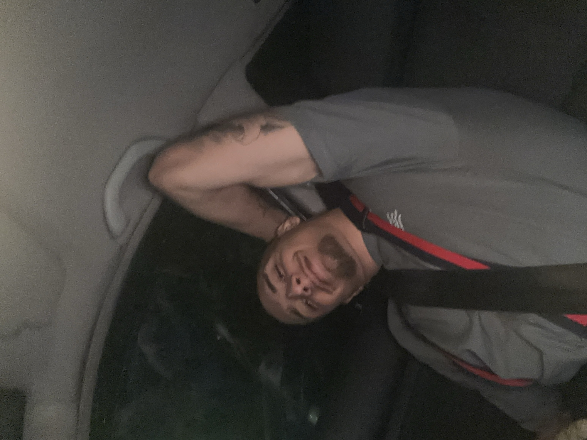 A man is lying down in what appears to be the back seat of a car. He is wearing a gray shirt with red stripes and has a tattoo on his right arm. He is wearing a seatbelt. It is dark inside the car. The specific location in Plovdiv, Bulgaria cannot be determined from the image.