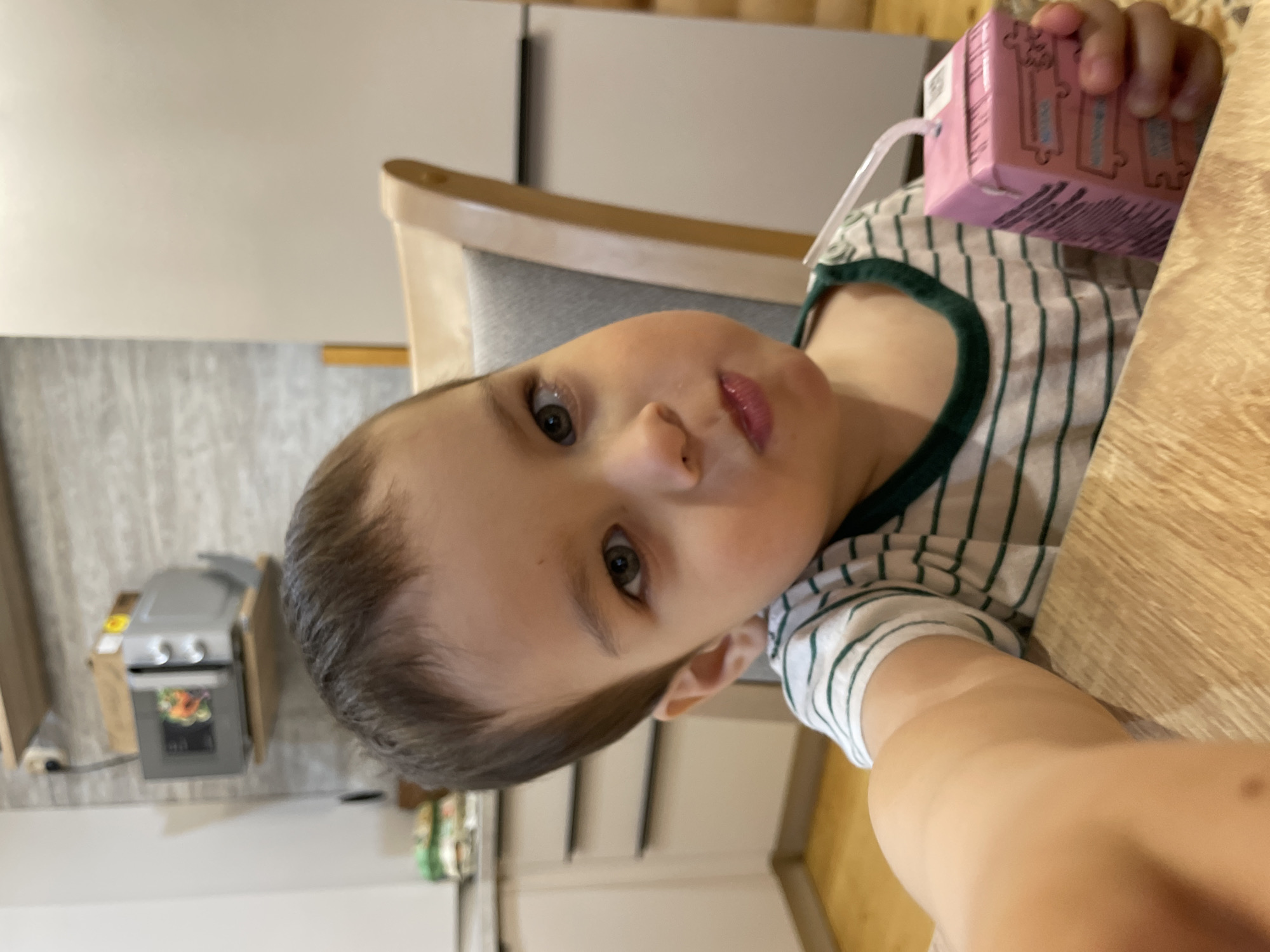 A young child is seated at a table, holding a pink juice box with a straw. The child has fair skin, short brown hair, and is wearing a white and green striped shirt. They are seated in a chair with a gray back and wooden frame. The table is wooden. In the background, a white refrigerator and a small oven are visible. This image does not provide enough context to determine its location within Zav'yalovo, Russia.