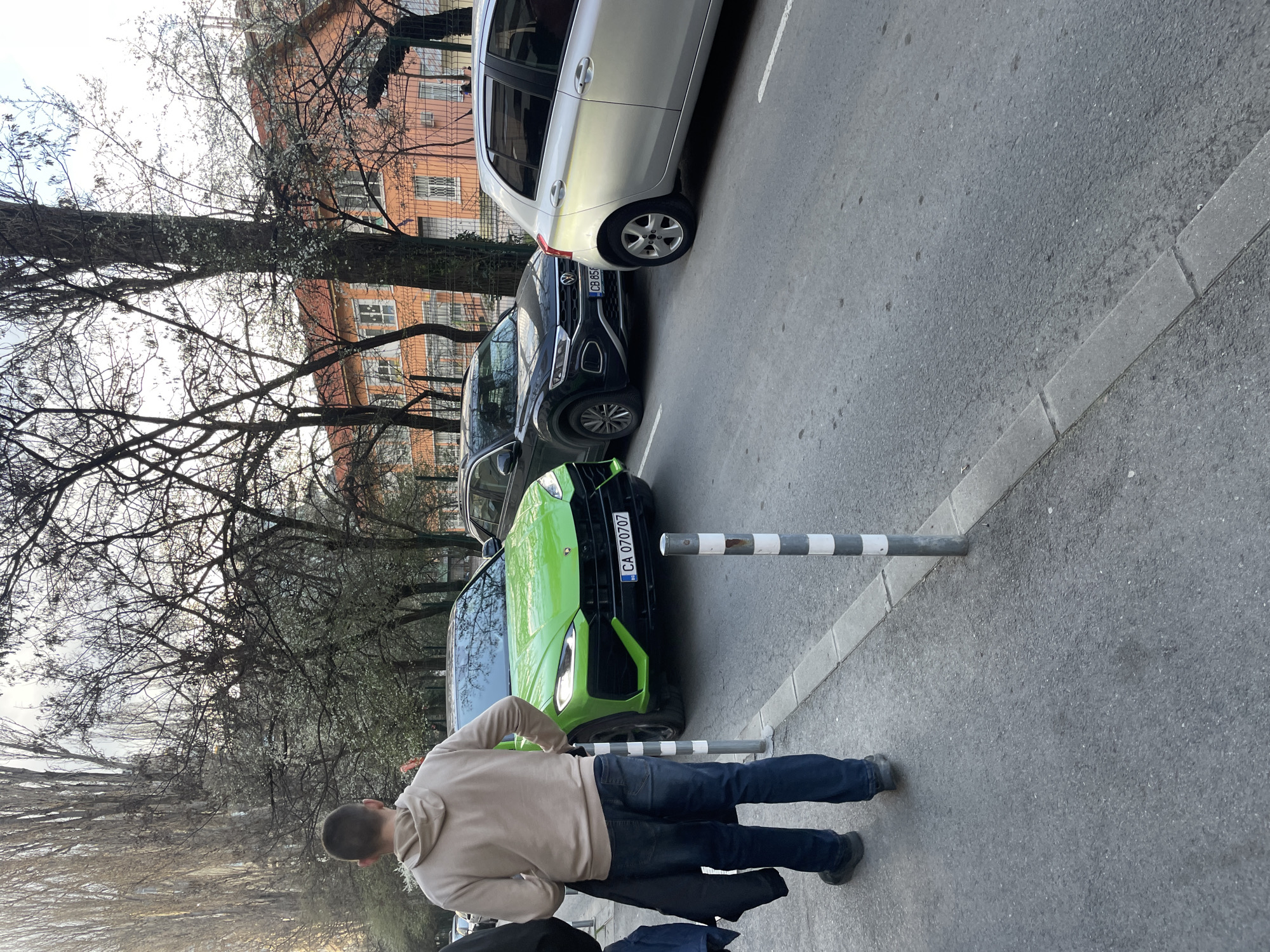 The image displays an urban street scene in Sofia, Bulgaria, likely during late afternoon or early evening in the spring. In the foreground, a male subject, viewed from behind, walks on a concrete sidewalk from left to right. He wears a light tan hooded sweatshirt, dark blue jeans, and dark shoes, carrying a dark jacket over his left arm. Black and white striped bollards are spaced along the sidewalk's edge. Three vehicles are parked parallel to the curb on the asphalt street. The prominent vehicle is a bright neon green Lamborghini Urus SUV, with its headlights illuminated, displaying Bulgarian license plate "CA 070707". Behind it, a dark Volkswagen T-Roc SUV is visible, showing a partial Bulgarian license plate "CB 85...". The third vehicle is a silver five-door hatchback. The background features several trees, some bare and tall, while others display nascent white blossoms. Behind the trees and a chain-link fence, a multi-story building with an orange facade, white-framed windows, and a reddish-orange tiled roof is visible.