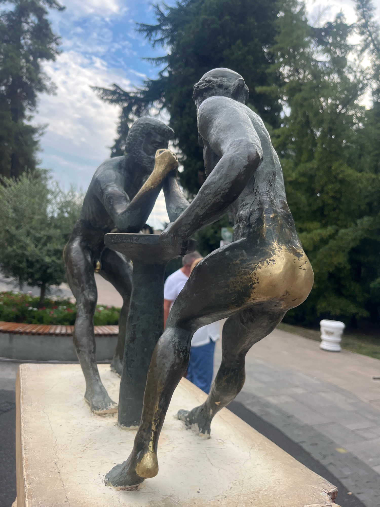 The image shows a bronze sculpture of two men arm wrestling, situated on a stone pedestal in a park setting. The sculpture is detailed, with the figures appearing muscular and engaged in the struggle. One figure's right hand and arm are gilded. In the background, lush green trees and a cloudy sky are visible. To the left, a wooden bench and some flowering bushes are present. A person in a white shirt and blue pants is partially visible behind the sculpture. To the right, a white cylindrical object and a paved pathway are seen. The sculpture is located in the Riviera Park in Sochi, Russia.