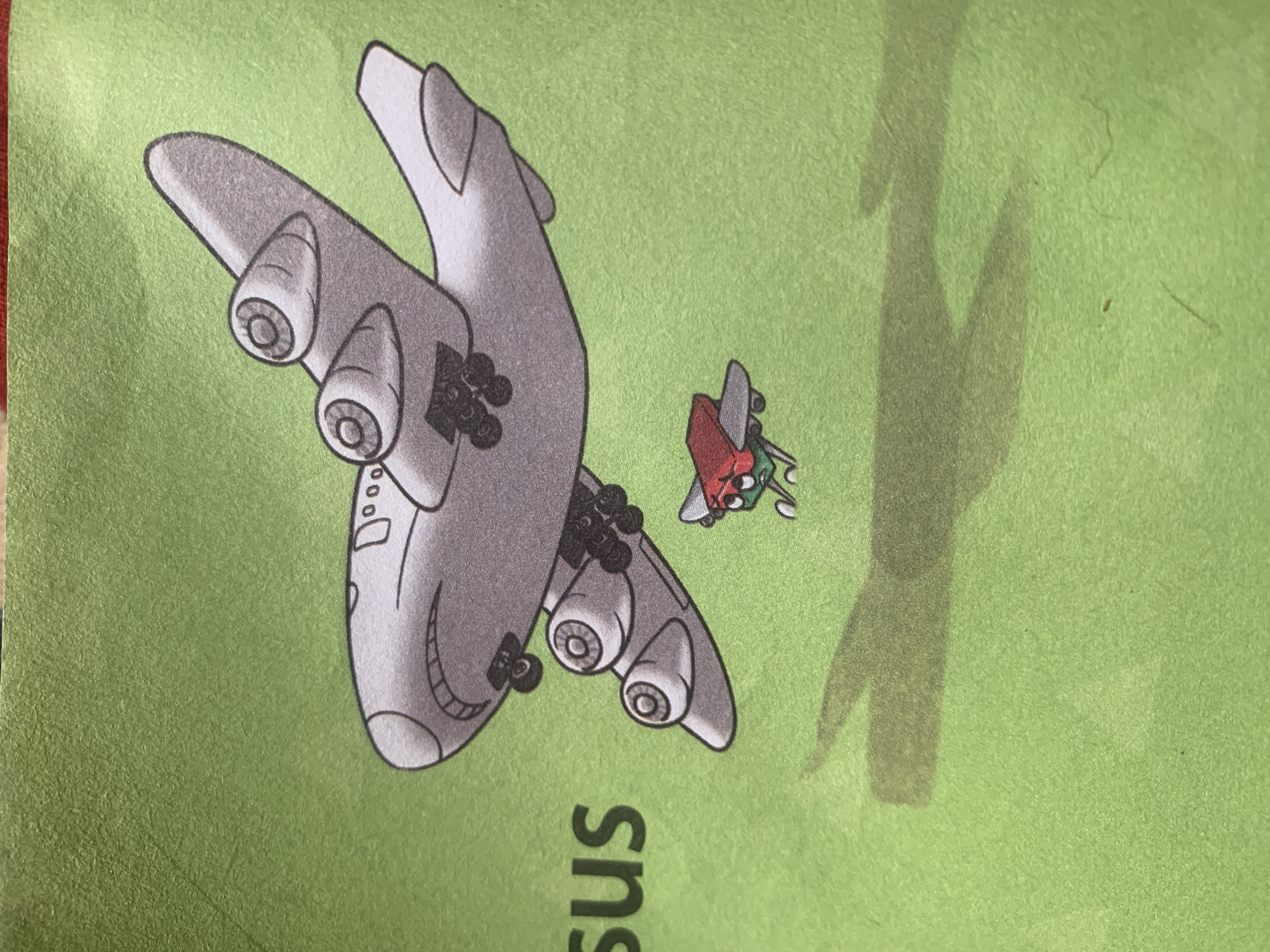 The image displays a cartoon illustration on a green background. The primary subject is a large, gray airplane with four engines, positioned in the upper right quadrant of the image. Below and to the left of the large airplane is a smaller, red and green airplane with a distressed expression. The word "sus" is visible in dark green text to the left of the large airplane. Shadows of both airplanes are cast on the green surface below them. The image does not depict any specific location in Jauro Yinu, Nigeria.