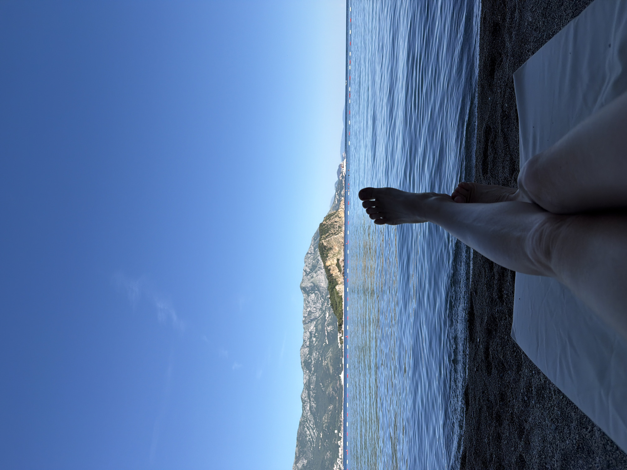 A view of the legs and feet of a fair-skinned person lying on a white cloth on a dark gravelly beach. Toes point toward the sea. The Adriatic Sea is characterized by blue, rippled water. A row of white and red buoys is visible further out. On the far side of the water is a coastal mountain range. The bottom of the mountains contains buildings of Budva, Montenegro. A clear blue sky fills the rest of the frame.