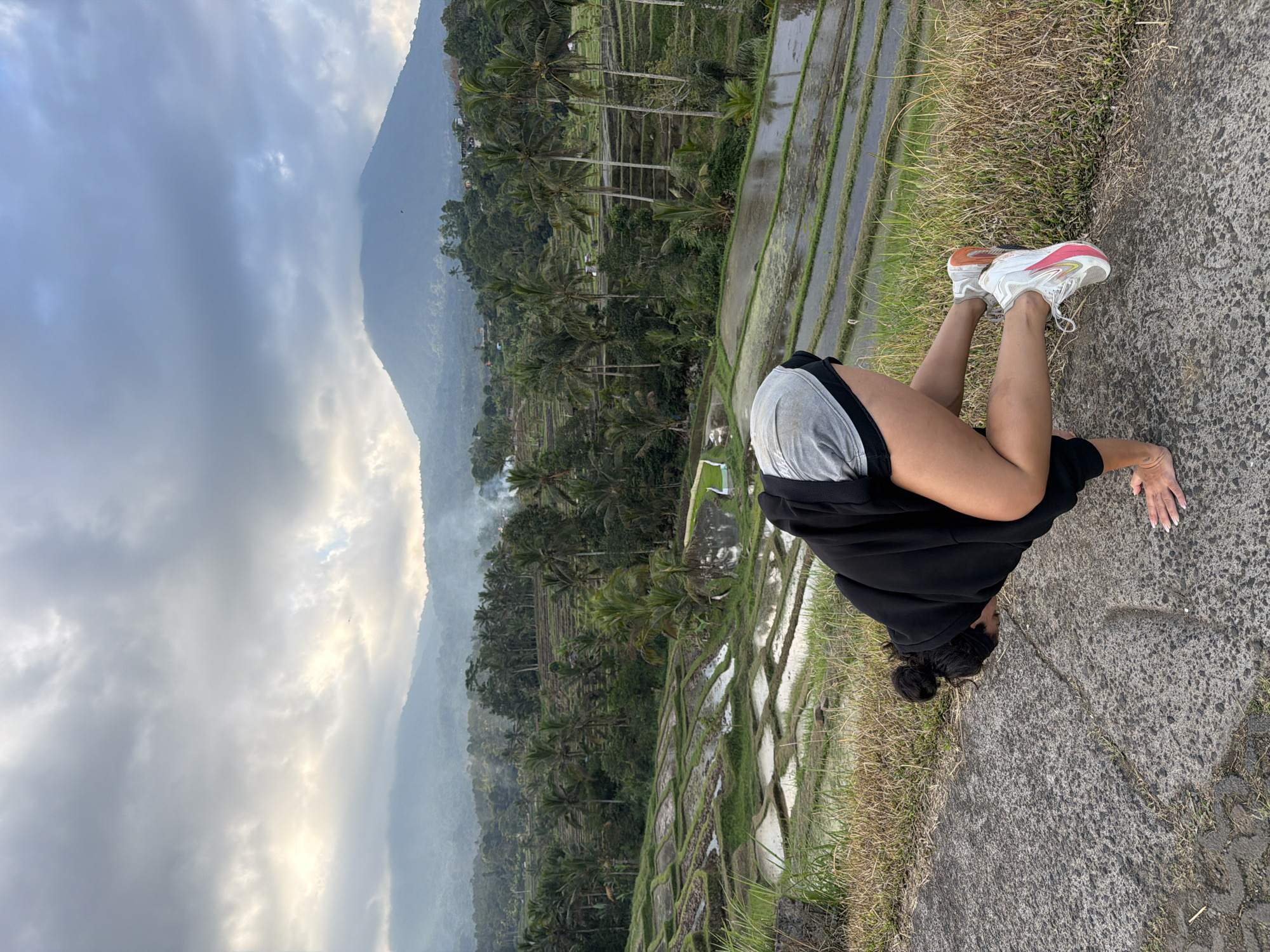 A person is performing a yoga pose on a paved surface with a backdrop of lush green rice terraces and a misty mountain range under a cloudy sky. The person is facing away from the camera, with their hands on the ground and their legs bent, balancing on their hands and one foot. They are wearing a black hoodie, gray shorts, and white sneakers with pink accents. The rice terraces are intricately carved into the hillside, with water reflecting the sky and surrounding vegetation. Smoke can be seen rising in the distance from a village nestled among the trees. The overall scene suggests a serene and natural environment in Penebel, Indonesia, a location known for its picturesque rice terraces.