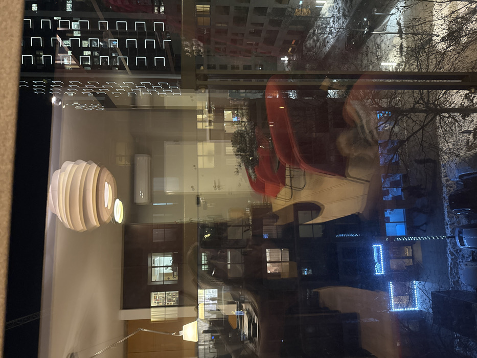 A complex window reflection shows a room interior with a modern light fixture and furniture, layered over an exterior scene of buildings at night, some with illuminated windows. A snow-covered ground and bare trees are visible. Christmas lights decorate one building. The reflection distorts the perspective and creates an abstract composition, blending indoor and outdoor elements.