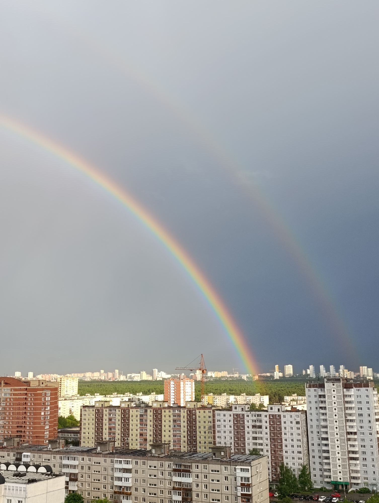 The image depicts a cityscape, likely in Perm, Russia, under a dramatic sky featuring a double rainbow. The foreground is dominated by Soviet-era apartment blocks of varying heights and colors (brick red, gray, off-white), each with numerous windows and balconies. A construction crane stands amongst the buildings, partially obscuring the view of trees in the midground. In the distance, a faint skyline of taller buildings is visible. The sky is overcast and divided: a lighter gray on the left side where the primary rainbow is prominent and a darker, stormy gray on the right where the secondary rainbow appears. The primary rainbow is brighter and more distinct than the secondary. No people are visible in the scene. The overall impression is of a residential area under changing weather conditions.