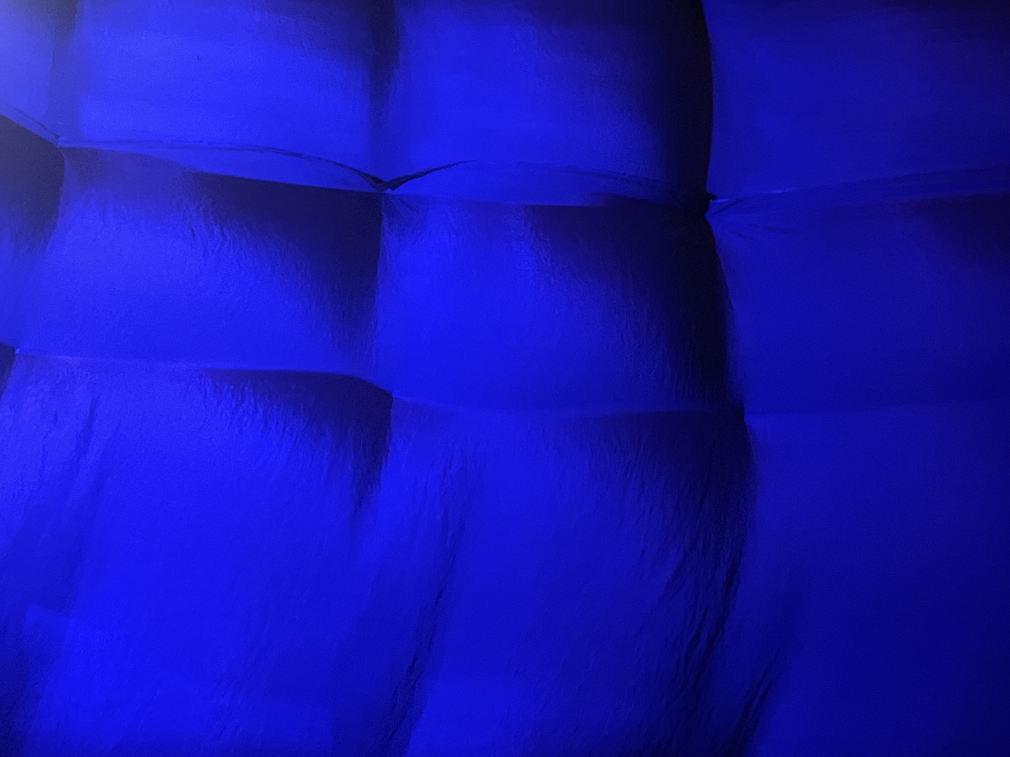 The image is a close-up, abstract shot of a dark blue, quilted surface. The texture appears to be fabric, possibly inflated or padded, with distinct sections creating a grid-like pattern. The lighting is dim and uniformly blue, casting soft shadows and highlighting the contours of the material. There are no discernible people, animals, or specific objects in the frame, creating a sense of depth and texture rather than a representational scene. The location in Mũi Né, Vietnam is not identifiable from the visual information provided in the image.