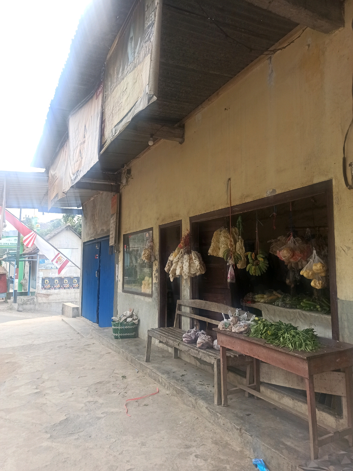 The image depicts an outdoor market scene in Tlogosari, Indonesia. The foreground shows a concrete ground with a red ribbon-like object on it. A wooden bench is placed in front of a building with a yellowish, weathered facade. A wooden table with legs is positioned in front of the bench, holding a bunch of green leafy vegetables.
Behind the bench, the building has an open doorway revealing a display of produce. Hanging from the top of the doorway are various food items, including bunches of bananas, dried snacks, and bags of produce. Inside the building, more vegetables are displayed on shelves.
To the left of the building, there is a blue double door and a window. Further to the left, flags are visible, including the Indonesian flag. The scene appears to be a small, informal marketplace. The overall environment is sunny, indicating daytime.