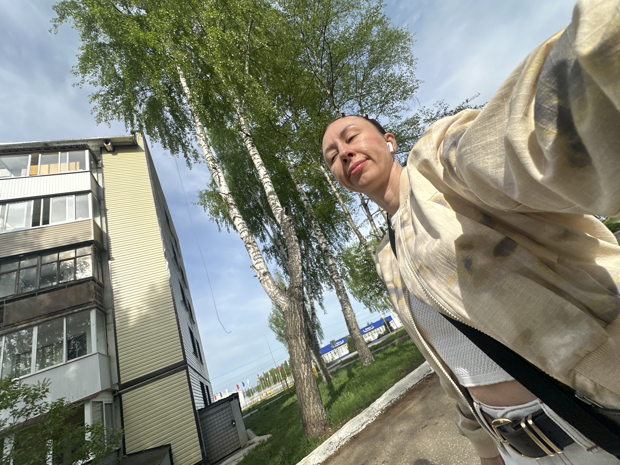 A selfie is taken outdoors in Votkinsk, Russia, capturing a woman wearing a beige patterned jacket, a white top, and a black belt with a gold buckle. She has an earbud in her ear and a neutral expression. The background shows a multi-story building with beige siding, a grassy area with birch trees, and a distant blue building. Flags are also visible in the distance. The sky is partly cloudy. The location is potentially near a park or recreational area based on the flags and trees.