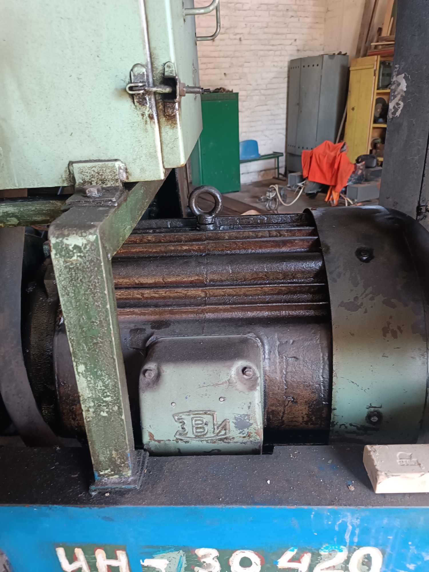 The image shows a close-up of industrial equipment, likely a motor, in what appears to be a workshop or storage area. The motor casing is dark and shows signs of wear and oil. A light green metal housing is attached to the top and left of the motor, also appearing weathered. In the background, there are various metal cabinets, shelves, and what looks like a piece of orange fabric. The environment suggests an industrial or manufacturing setting. The floor is dark and appears to be concrete or a similar material. The image is well-lit, highlighting the textures and details of the machinery. There is no indication of the specific location within Vuhlehirsk, Ukraine, beyond it being an industrial setting.