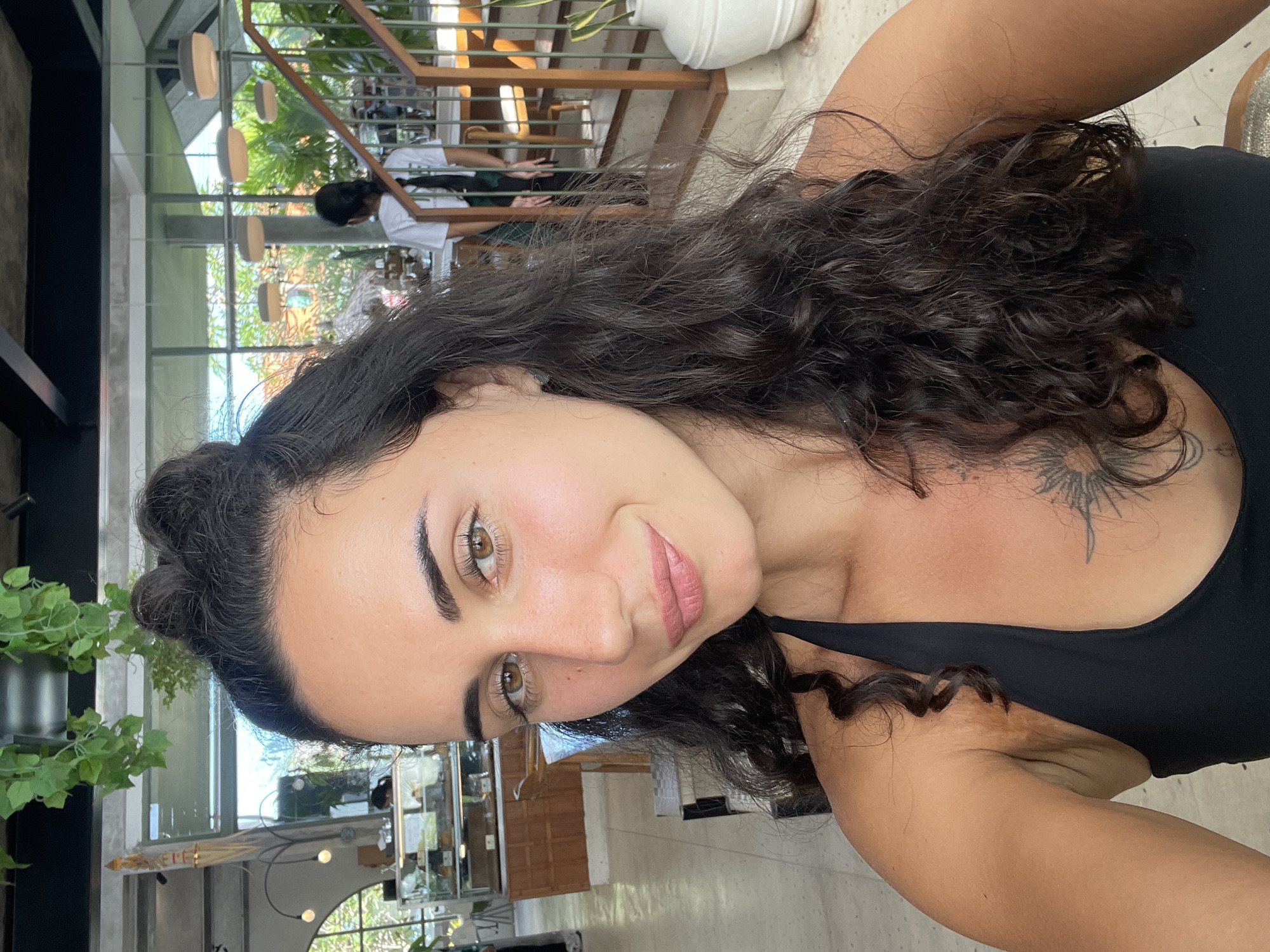 A woman with dark curly hair and brown eyes is taking a selfie. She is wearing a black tank top. A small tattoo is visible on her upper chest. The background appears to be an indoor/outdoor space, possibly a restaurant or cafe. The setting is bright and airy, with natural light filtering through windows. Vegetation and architectural elements suggest the location is in North Kuta, Indonesia. Several people are visible in the background.
