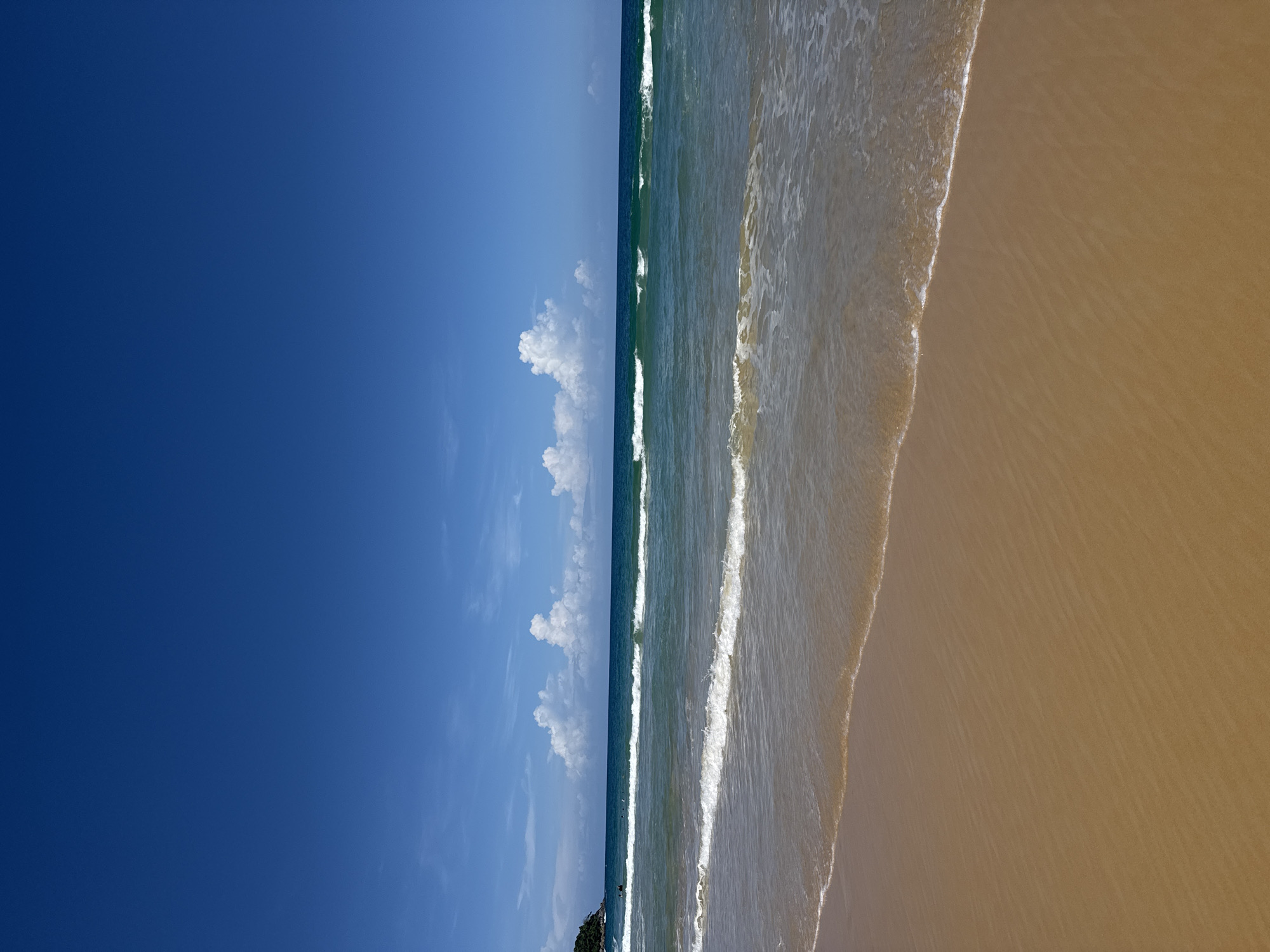 The image depicts a serene beach scene at Karon Beach in Karon, Thailand. The foreground is dominated by a stretch of golden sand, marked with subtle ripples from the receding tide. A gentle wave, with a foamy white crest, is making its way towards the shore, partially obscuring the sand. Beyond the immediate shoreline, the clear turquoise water of the Andaman Sea stretches towards the horizon, with several smaller waves creating patterns on its surface. In the distance, the deep blue sea meets a bright blue sky, dotted with fluffy white cumulus clouds. A faint silhouette of a lush green headland can be seen on the far left of the image, hinting at the edge of the beach. A few people are visible in the water, appearing as small dark figures enjoying the ocean. The overall atmosphere is tranquil and idyllic, characteristic of a tropical beach destination.
