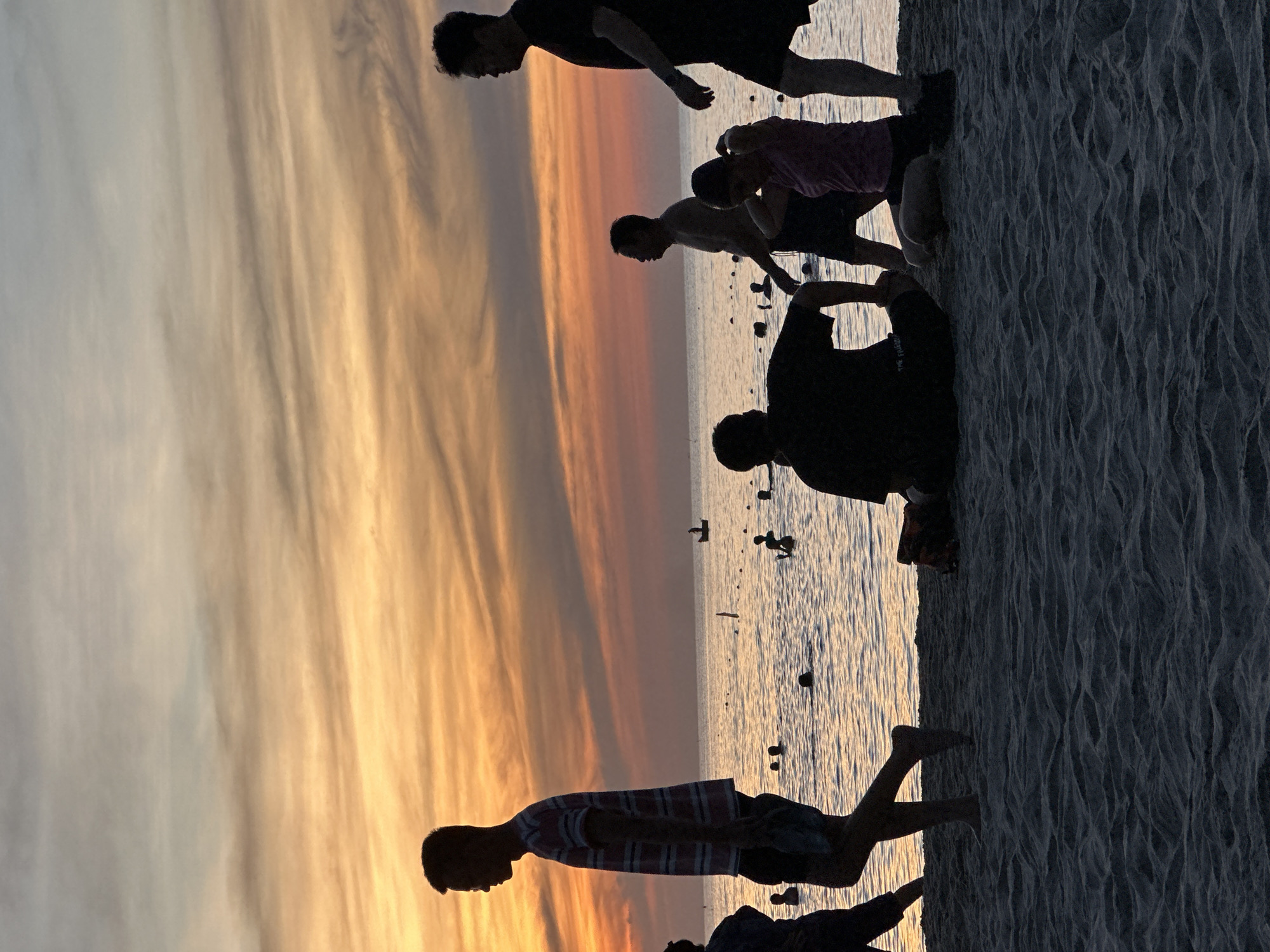 At sunset on a beach in Đà Nẵng, Vietnam, the silhouettes of people are visible against a vibrant orange and pink sky. In the foreground, several individuals are on the sand, some walking and others sitting. A man in a striped shirt walks from left to right, while another man sits with his back to the camera. A woman in a pink top is kneeling in the sand. Further back, more people are visible on the beach and in the shallow water, some swimming. A small boat is also visible on the calm sea. The sand in the foreground shows the texture of waves from the receding tide.