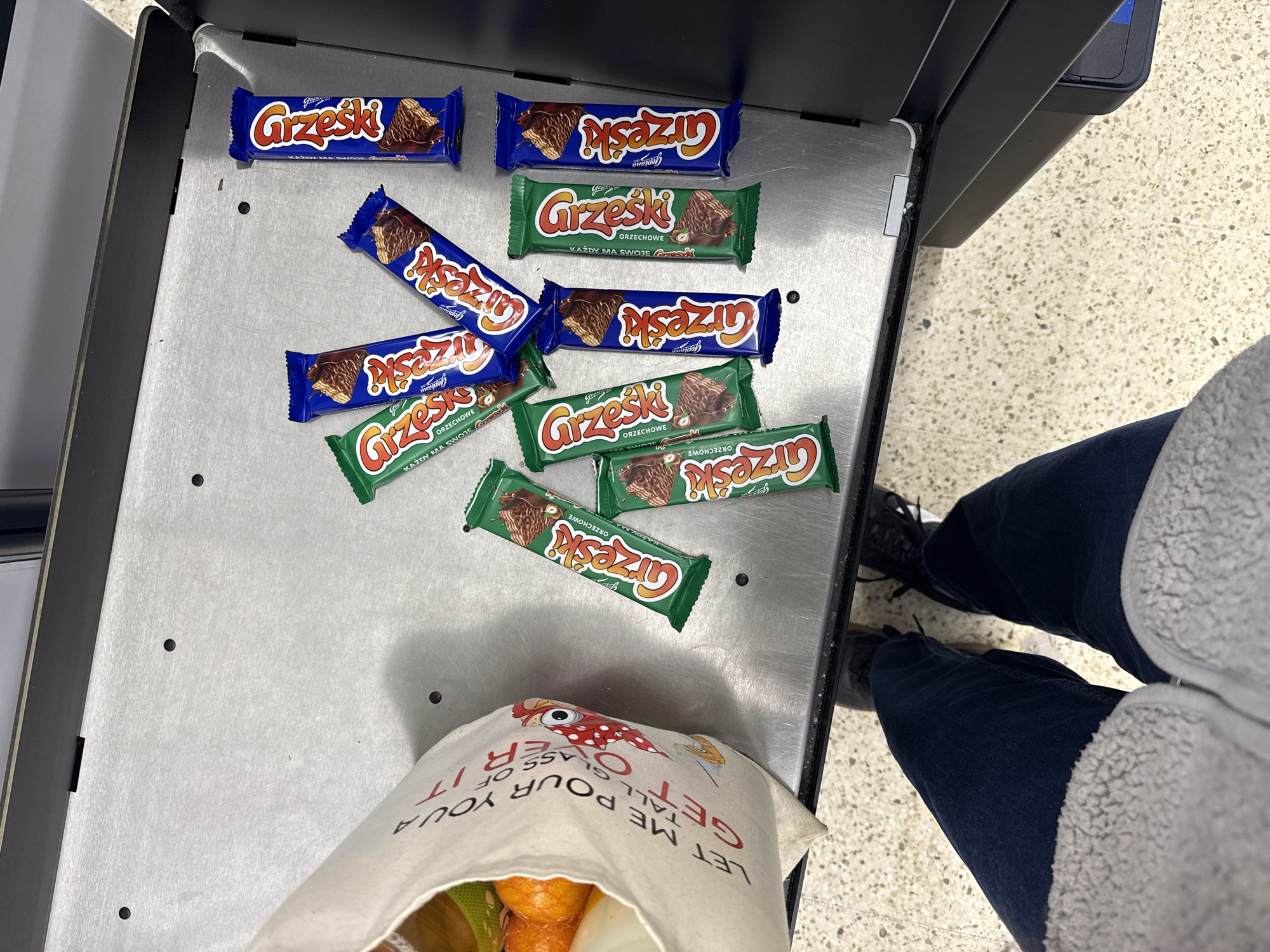 A high-angle, medium shot shows an assortment of "Grzeski" wafer candy bars on a metallic checkout conveyor belt. There are five blue-wrapped and six green-wrapped candy bars. Below the candy bars, a partially filled beige tote bag rests with its contents partially visible, including oranges and a green-and-yellow object. The bag has a cartoonish red-and-white mushroom graphic and the text "LET ME POUR YOUR TALL GLASS OF IT / GET OVER." The person operating the checkout is visible from the knees down, wearing dark-wash jeans and a light gray sweater, with black shoes visible. The location is not specified, however, the OCR contains a reference to Goplana, a polish confectionary company that produces the "Grzeski" bars. The city of Great Yarmouth is not discernible from the image.