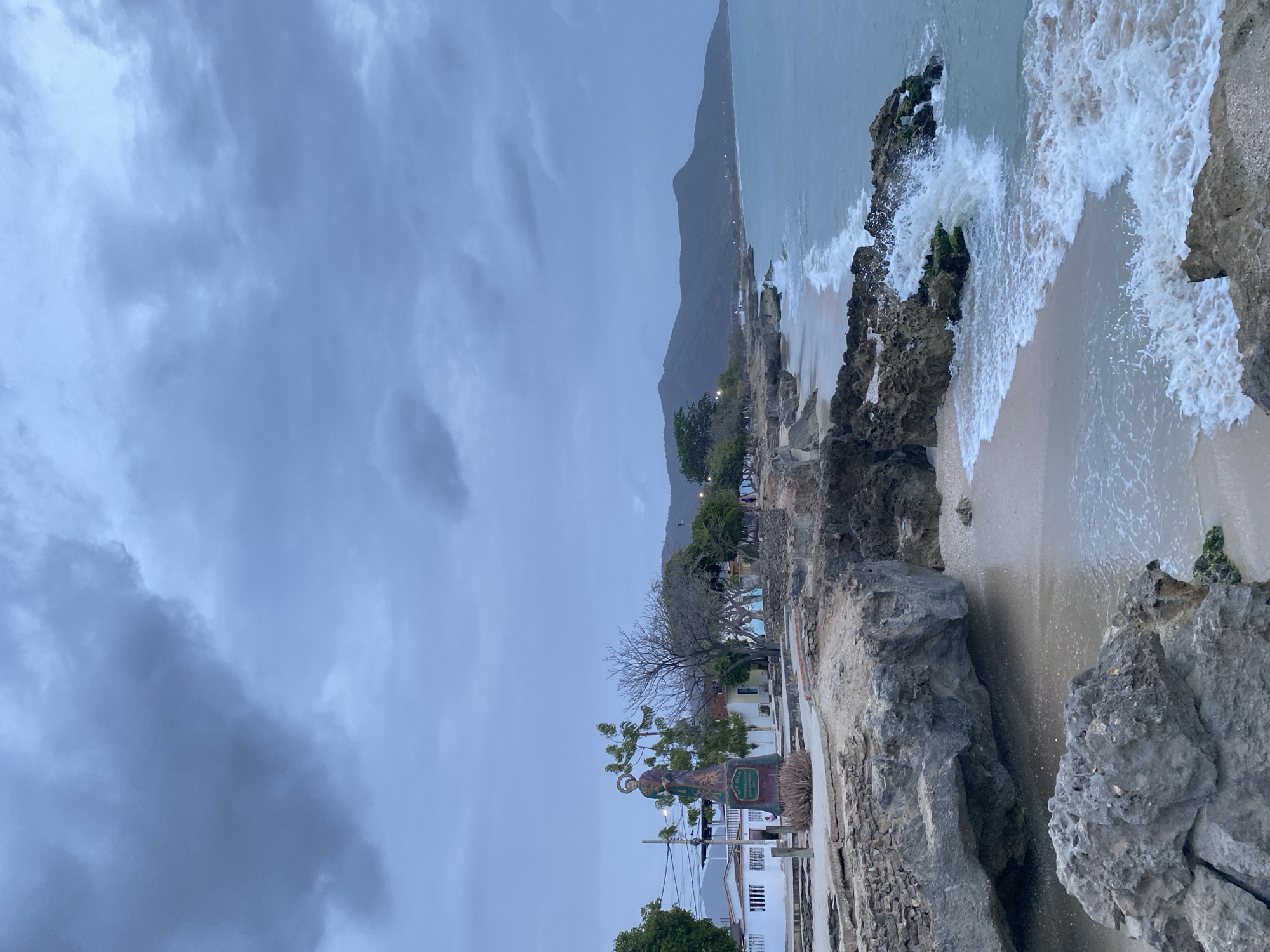 The photograph captures a coastal scene, likely in Juan Griego, Venezuela. The sky is overcast with heavy, gray clouds, suggesting a recent or impending storm. A mountain range rises in the background. In the foreground, rocky outcroppings meet the shoreline, where waves crash against the rocks and beach. A strip of sandy beach is visible. Adjacent to the rocky coast, there is a paved area with buildings, trees, and a statue of a figure dressed in gold and green. In the middle ground, there are buildings, trees and light poles.