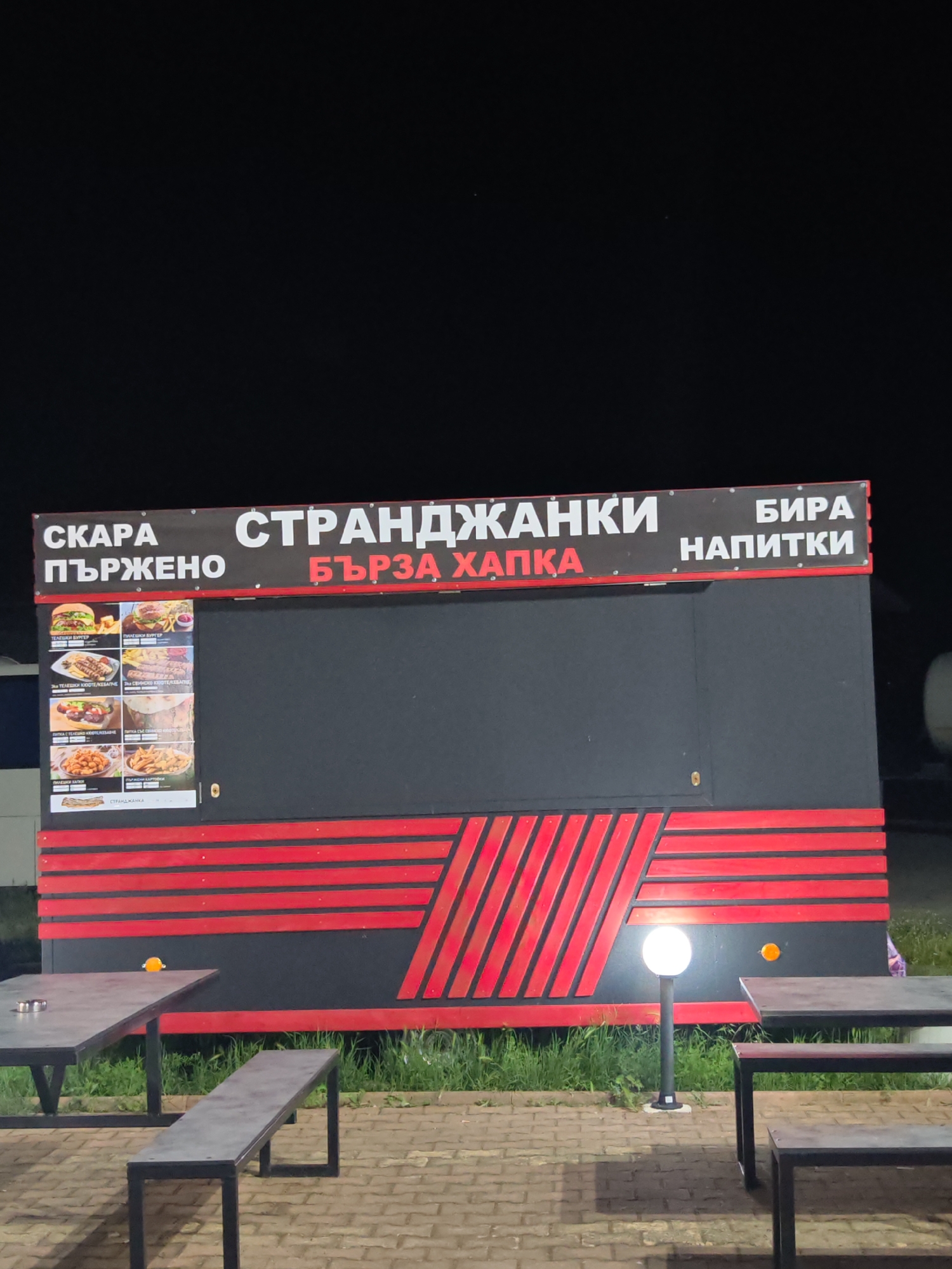 At night in Yabalchevo, Bulgaria, a food stand named "Strandjanki" (presumably a local specialty) is visible. The stand has a black exterior with red horizontal stripes and angled red lines. The sign above it advertises "Skara Purjeno" (Grilled Fried), "Barza Hapka" (Quick Bite), and "Bira Napitki" (Beer Drinks). A menu with images of food items is also displayed. In front of the stand are tables and benches. A street light illuminates the area. The ground is paved. Grass is visible between the pavement and the stand. The sky is dark.