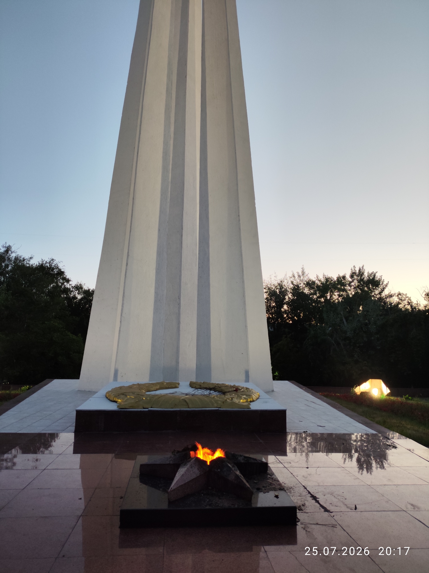 The image shows a memorial monument with an eternal flame in the foreground. The monument is a tall, white, fluted obelisk. In front of the obelisk is a wreath made of gold-colored material. The eternal flame burns in a star-shaped receptacle. The ground around the memorial is paved with polished, dark red granite tiles that reflect the monument and the flame. In the background, there are trees and a lit tent. The sky is a pale blue, suggesting dusk or dawn. The date and time "25.07.2026 20:17" are visible in the bottom right corner. This memorial is the Eternal Flame at the Eternal Flame Memorial Complex in the city of Pavlodar, Kazakhstan, dedicated to fallen soldiers.