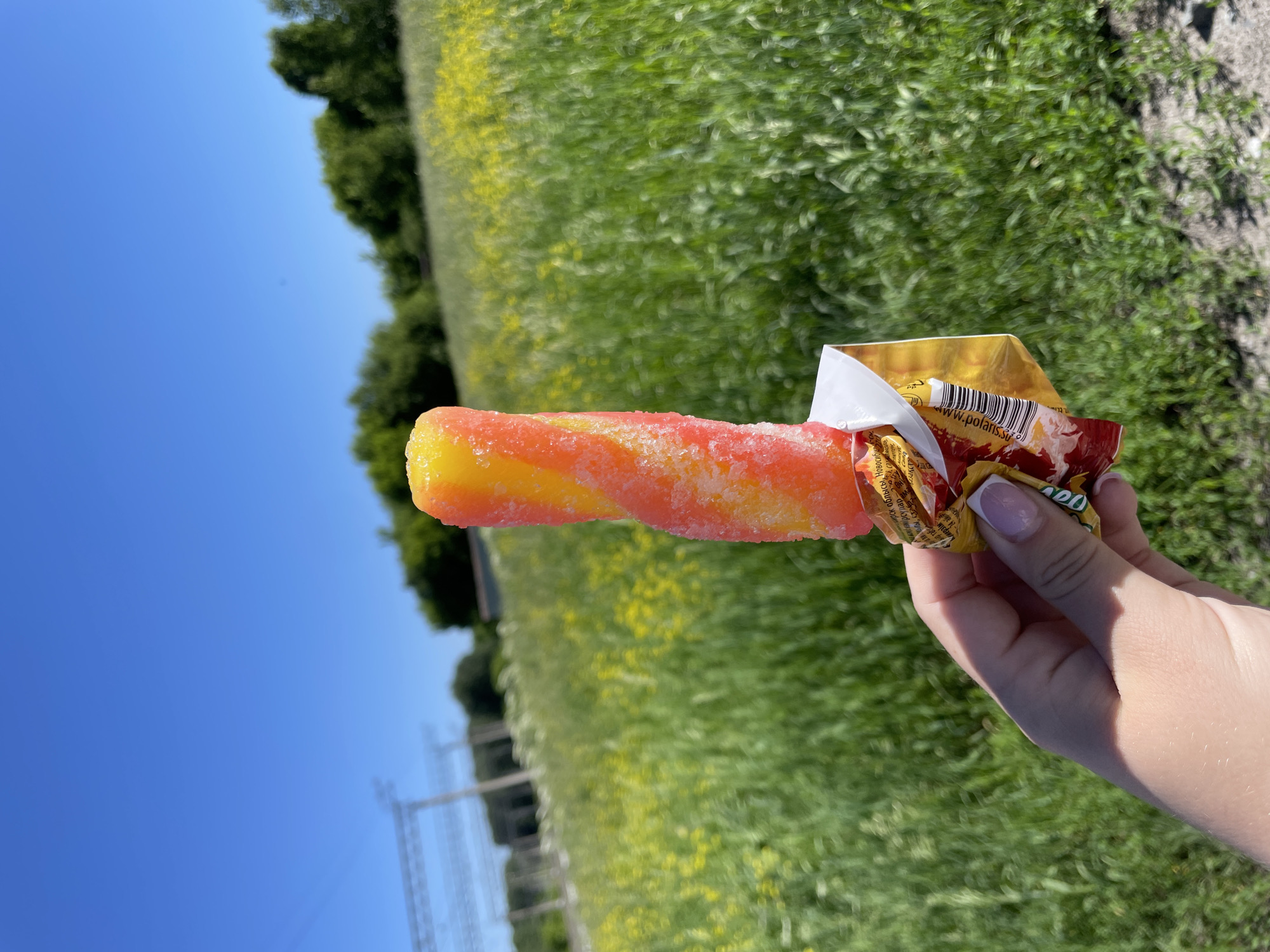 In Artyshta, Russia, a hand with a pink-tipped manicure holds a vibrant orange and yellow popsicle. The popsicle is partially unwrapped from its red and yellow packaging, with visible text on the wrapper. The background features a bright blue sky and a field of green grass dotted with yellow flowers. In the distance, trees form a treeline, and a set of power lines is visible on the left. The overall scene depicts an outdoor summer day.