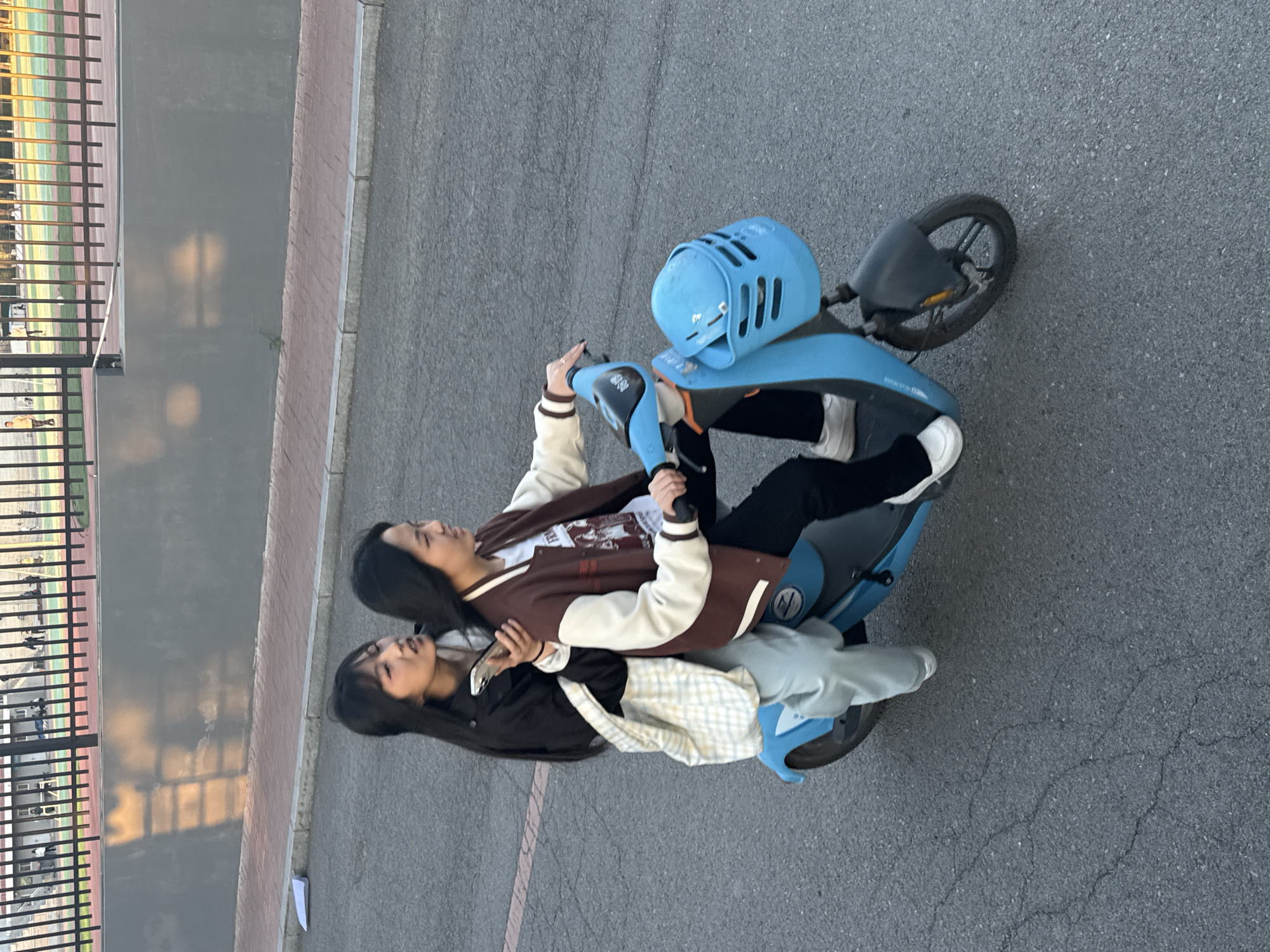 Two young women are riding a blue electric scooter on a gray asphalt surface, possibly a street or parking lot. The woman in front is wearing a brown and white varsity jacket with black pants and white sneakers. She is holding onto the handlebars. The woman behind her is wearing a black jacket with a white and gray checkered shirt. Her left hand is holding a smartphone. A metal fence is visible in the background, with a brick sidewalk adjacent to it. The scene appears to be outside, likely in Zhongtun, China.