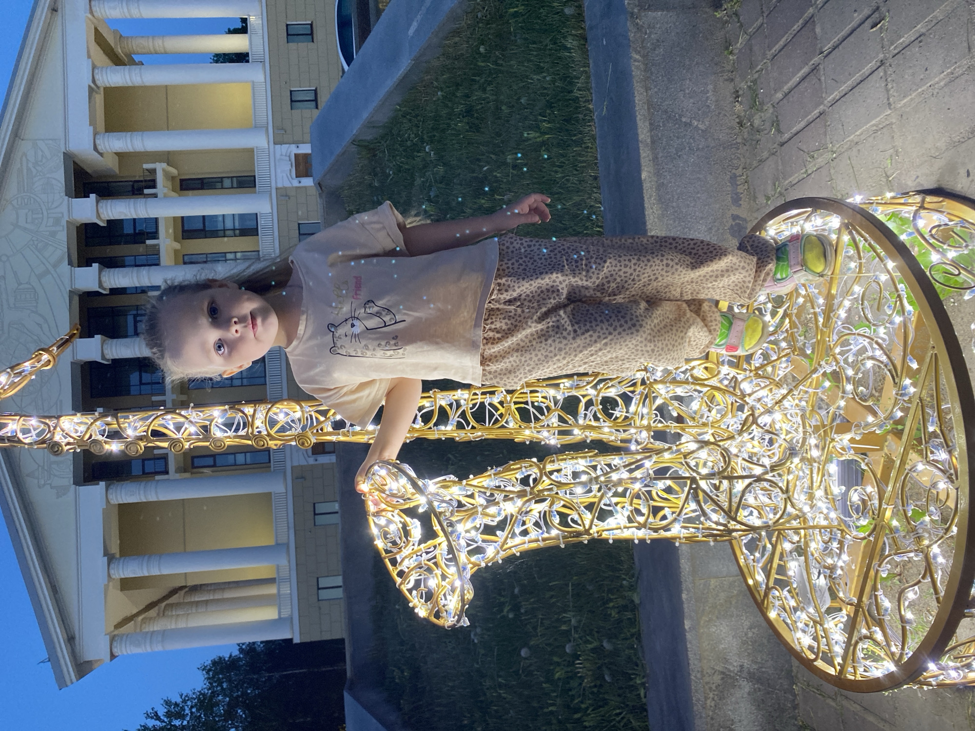 A young girl is photographed standing next to a decorative light structure in what appears to be the city of Votkinsk, Russia. The girl wears a light-colored shirt with a cat graphic and leopard-print pants, and multicolored sneakers. She stands on the light structure's base, holding onto part of the metal frame. The decorative light structure is gold and illuminated with small lights, possibly shaped like a reindeer or a horse, with a circular base. In the background is a building with white columns and windows, suggesting a public building or monument. To the side, there's a patch of grass and a paved area, suggesting an outdoor setting, possibly a park or public square. The sky suggests that it is evening, with a transition from light to dark blue.