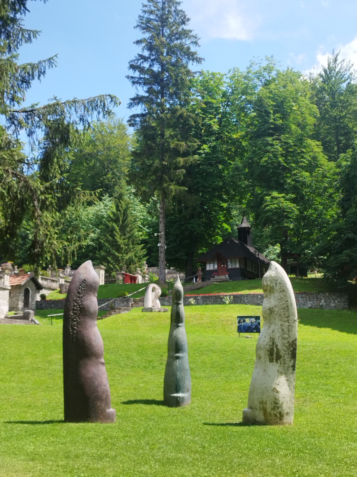 The image depicts an outdoor art installation in the city of Bușteni, Romania, specifically within the Cantacuzino Castle gardens. In the foreground, three abstract stone sculptures stand on a lush green lawn. The sculpture on the left is dark brown with a textured surface, appearing to be made of bronze or a similar dark metal. The central sculpture is slender and dark green, with a smooth, tapered form. The sculpture on the right is lighter in color, appearing weathered and perhaps made of a lighter stone or concrete. In the middle ground, a small, dark wooden building resembling a chapel or pavilion with a steeple is visible. Stone staircases and retaining walls lead up a gentle slope behind the chapel, surrounded by dense green trees. A framed picture displaying three individuals in formal attire is placed on the lawn in front of the chapel. The sky is clear blue with a few scattered white clouds, and sunlight casts shadows on the grass. The overall impression is a serene and artistic park setting.