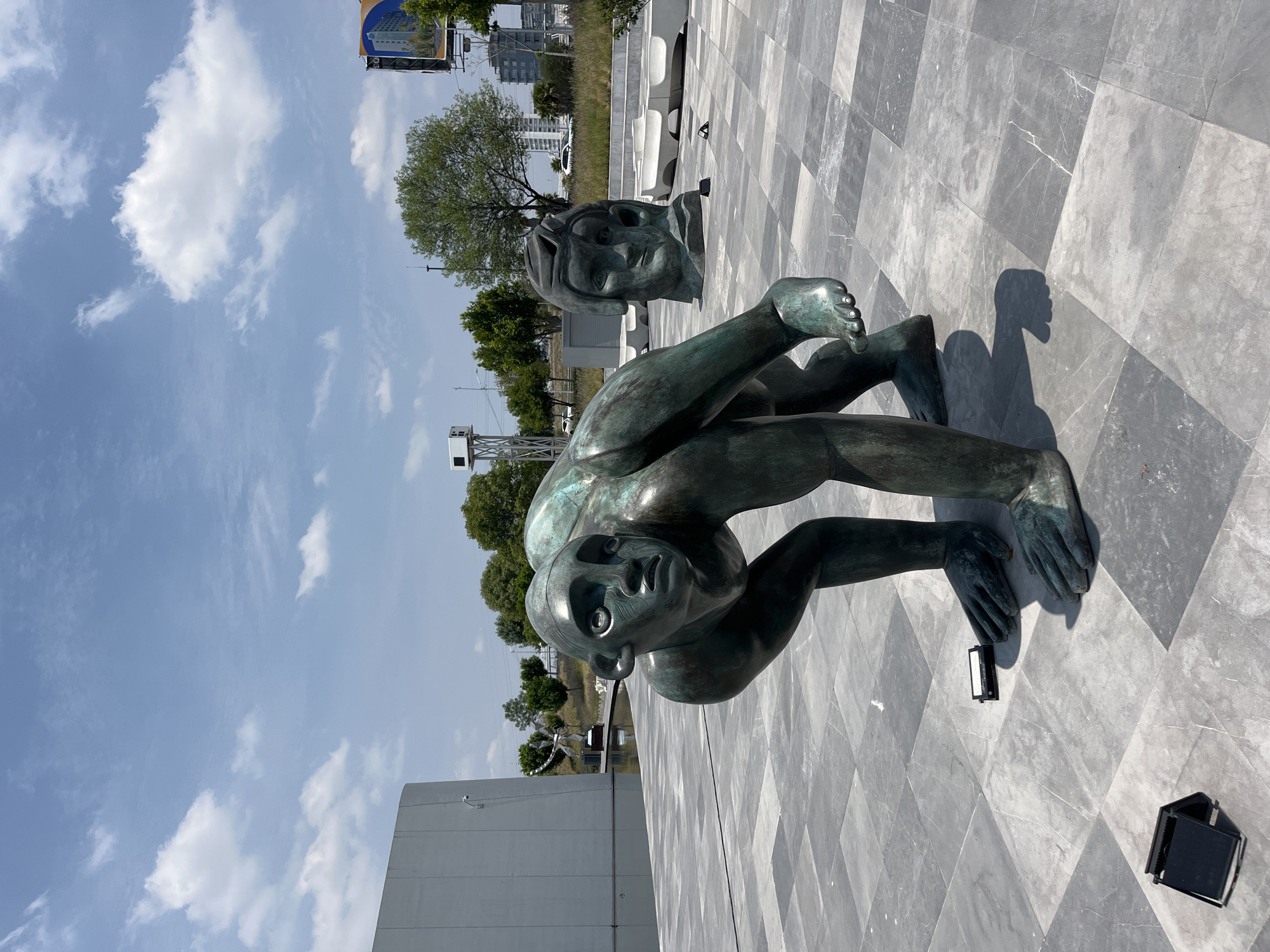 A ground-level outdoor shot in San Bernardino Tlaxcalancingo, Mexico, displays a bronze statue of a crouching human figure with a second human head sculpture placed behind it. The statue and head rest on a gray, diamond-patterned stone surface. The figure is crouched with arms extended toward the ground. The statue's features are simplified.
In the background, trees, a white tower structure, and buildings are visible under a blue, partly cloudy sky. A white building edge is partially visible in the lower left corner of the frame. Two black light fixtures are placed near the base of the statue. The scene is in daylight.