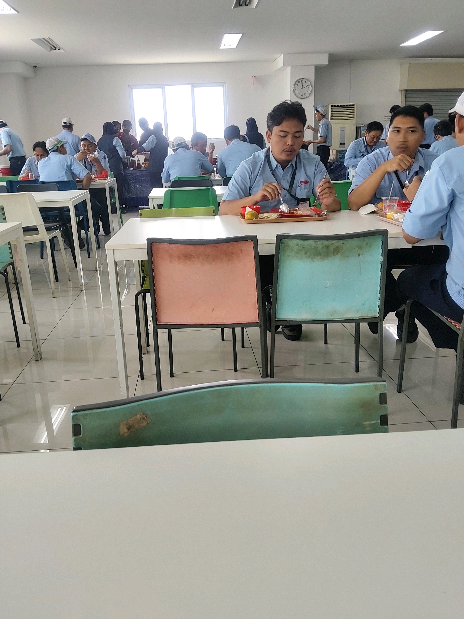 The image depicts a brightly lit indoor space that appears to be a cafeteria or dining hall, likely located in Mekarmukti, Indonesia, given the context of its origin. The environment is characterized by white tiled flooring and a white ceiling with overhead lighting panels. Several tables are arranged throughout the space, with people seated and eating.
In the foreground, a white table is prominently featured. Beyond it, a teal-colored chair with a worn, textured surface is visible. Further back, another table has two young men seated, both wearing light blue polo shirts with a red and white logo on the left breast. The man on the left is eating from a red plastic tray with a fork and spoon, while the man on the right is also eating from a tray. Several other individuals are seated at tables in the background, some engaged in eating and others in conversation. The overall atmosphere suggests a communal mealtime. The background includes windows letting in natural light and some industrial-looking equipment, possibly an air conditioning unit.