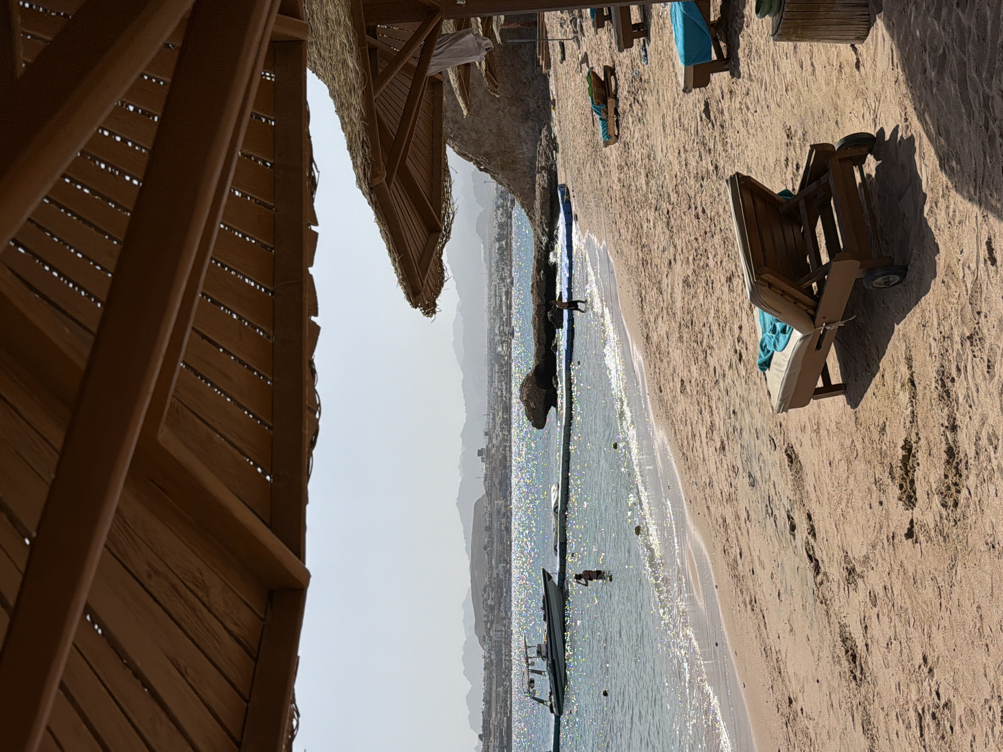 The image shows a beach scene in Sharm El Sheikh, Egypt. The viewpoint is from beneath a wooden thatched-roof structure, possibly a beach umbrella or cabana. Adjacent to this structure and the beach are a line of beach umbrellas, each providing shade over a sunbed. The beach slopes down to a shoreline where clear, light blue water meets the sand. In the water, there is a boat and a person standing, partially submerged. In the background, the city of Sharm El Sheikh stretches along the coastline, with mountains rising in the distance. The sky above the city is a clear, light blue, suggesting a sunny day.
