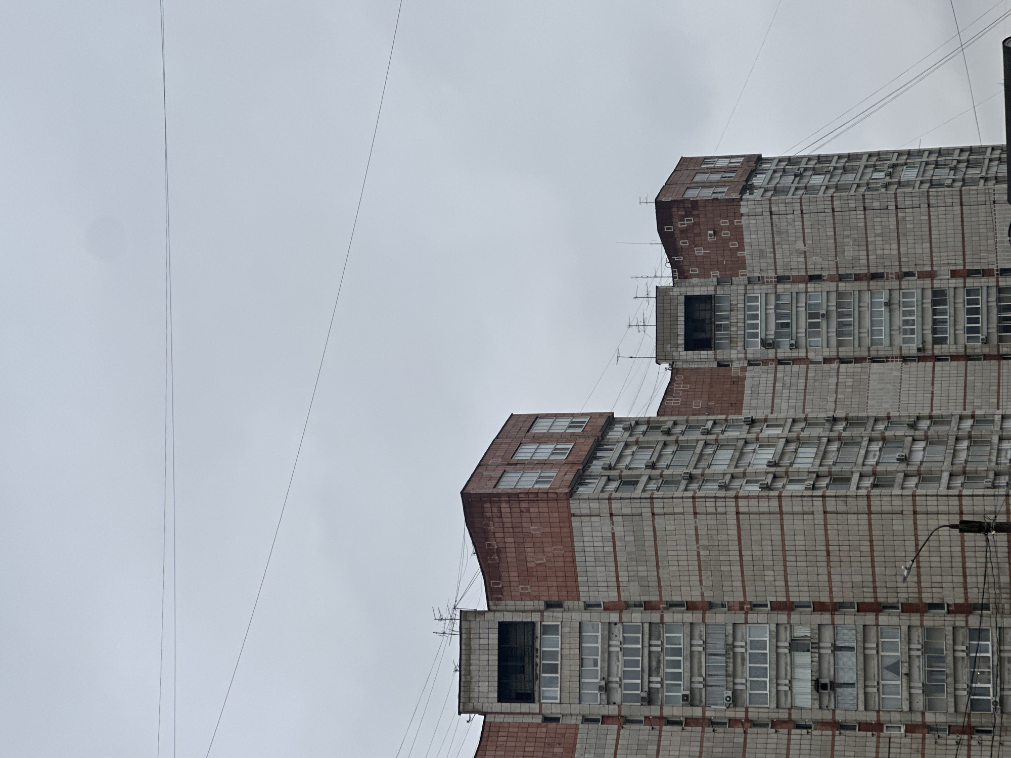 The image shows a multi-story building in Perm, Russia, characterized by a stacked design with receding upper sections. The building has a facade of gray and red tiles, punctuated by rows of rectangular windows. Multiple thin power lines are visible stretching across the overcast sky. A street light is mounted on a pole to the right of the building.