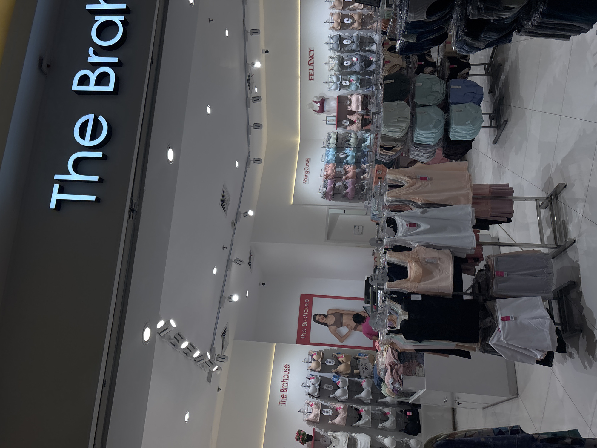 The image displays the interior of a well-lit intimate wear store, likely situated in a shopping center in North Jakarta, Indonesia. The store's name, "The Brahouse," is prominently featured in large, illuminated letters on the left wall. A large promotional poster further inside also shows "The Brahouse" branding with a woman modelling a bra, with a partially visible person in a pink top standing near it, browsing.
The store is meticulously organized, showcasing a wide range of intimate apparel, including bras, underwear, and basic tops. These items are displayed on multiple metal racks and torso mannequins along the right side of the store. The merchandise comes in various styles and colors, predominantly neutral tones like nude, white, and black, alongside pastels such as pinks, blues, and greens. Folded garments, possibly underwear or basic tops, are neatly arranged on shelves beneath the hanging displays.
Visible brand names on the walls include "Young Curves" and "FELANCY," indicating different collections or labels offered. A "DISCOUNT 70%" sign is clearly visible on one of the bra displays, advertising a sale. The store features a modern and bright aesthetic with abundant artificial lighting from recessed ceiling lights and track lighting, illuminating the white walls and light-colored tiled floor.