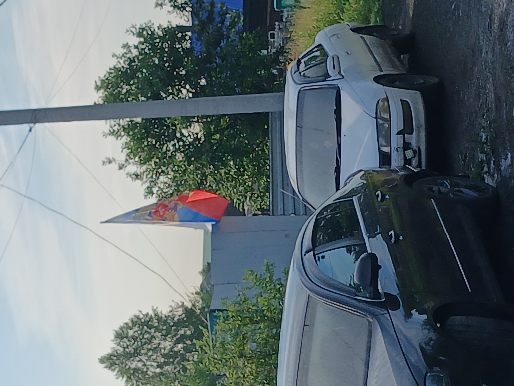 In Prokopyevsk, Russia, on a bright, clear day, two cars are parked on a dirt road. A dark-colored sedan is on the left, and a white sedan is on the right. Behind the cars, a grey concrete fence stands, partially obscured by green foliage. A pole, likely for power lines, is visible to the right of the fence. A flag, predominantly red, white, and blue with a crest, is attached to a wire and flutters in the breeze above the fence. The background consists of green trees and several houses with blue roofs. The scene appears to be a residential area in Prokopyevsk.