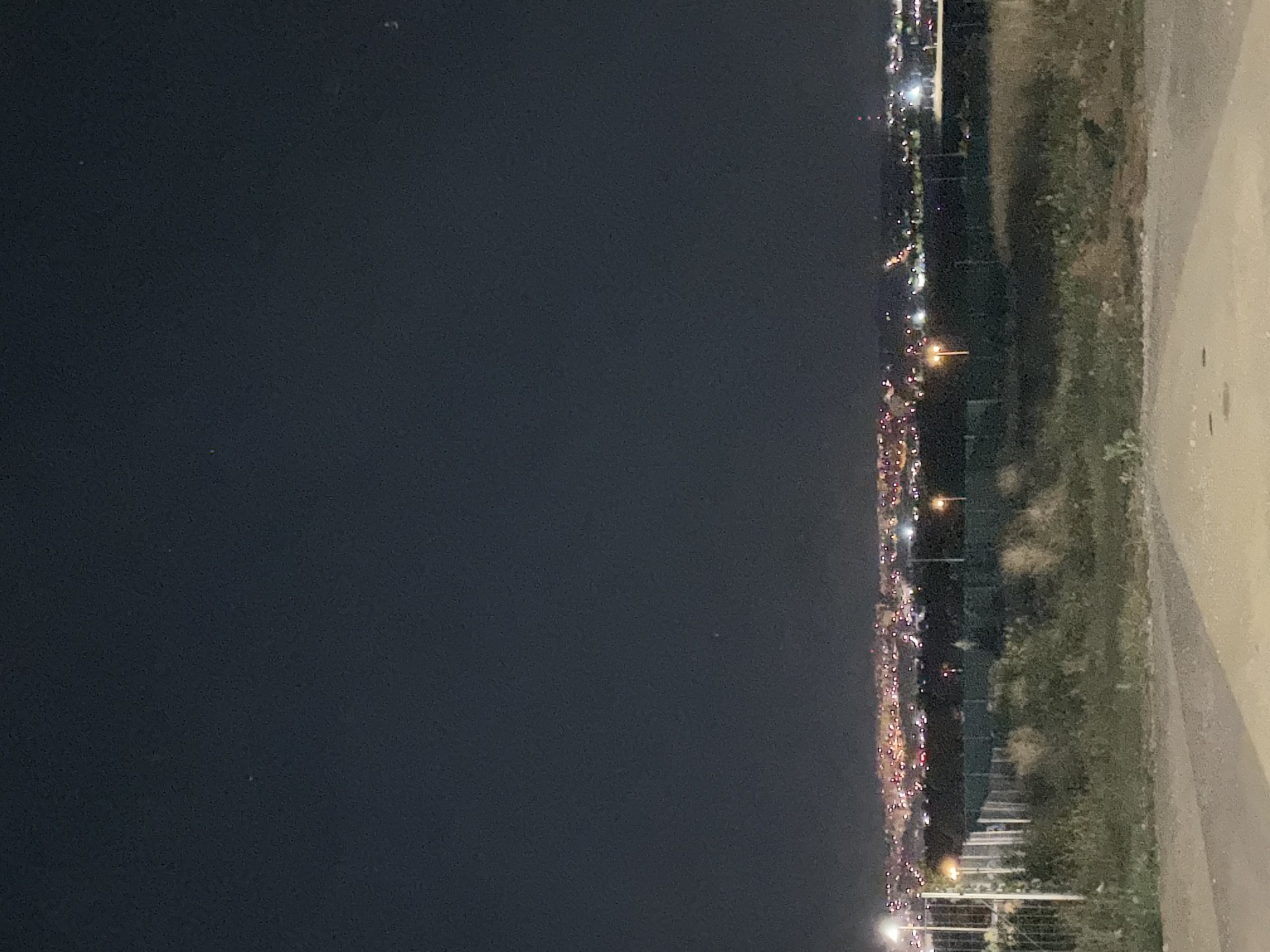The image shows a night view of a city with a dark sky above and a foreground of rough terrain and vegetation. In the mid-ground, there is a fence line with a few streetlights. Beyond the fence, a city skyline is visible with numerous lights from buildings and streetlights, indicating an urban area. The lights suggest a settlement spread across a landscape, possibly with some elevation changes given the city lights appearing to ascend. There is no specific identifiable landmark or feature that directly indicates the city is Nitra, Slovakia. The environment appears to be an elevated viewpoint overlooking the city at night.