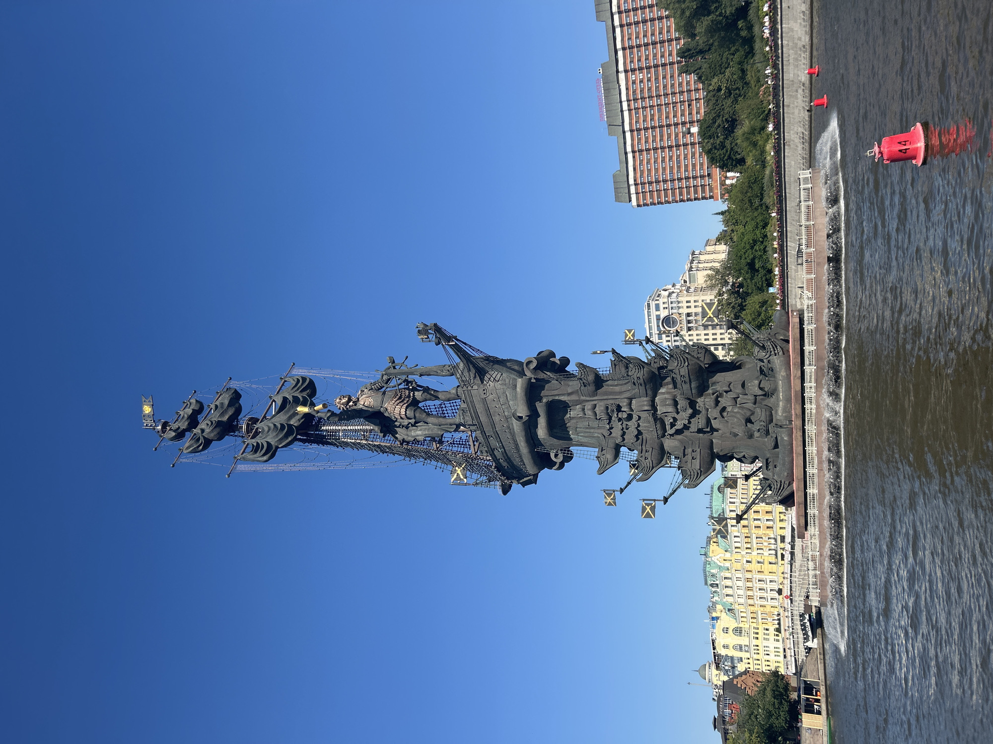 A large, bronze statue of Peter the Great atop a ship is situated on a fountain in the Moskva River in Moscow, Russia. The statue, a prominent landmark, depicts Peter the Great standing at the helm of a multi-decked vessel, adorned with flags and intricate rigging. Water sprays from the fountain surrounding the base of the statue, creating a dramatic effect. In the background, several multi-story buildings line the riverbank, including a large, modern hotel with a brick facade and a row of older, colorful buildings. Lush green trees are visible along the embankment, and the sky is a clear, vibrant blue. Three red buoys are floating in the river in the foreground.