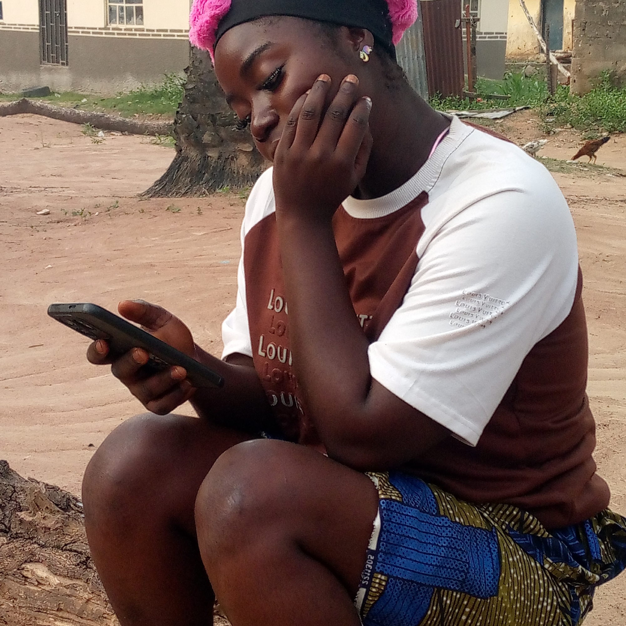 A young woman is seated outdoors, likely in Mararaba, Nigeria. She is dark-skinned with a pink headscarf and is engrossed in her phone, which she holds in her left hand. Her right hand is touching her face. She wears a two-tone shirt with "Louis Vuitton" repeated on the sleeves and a colorful blue and patterned fabric wrapped around her waist. She sits on what appears to be a weathered tree stump. In the background, there is a building and a chicken walking on the dirt ground.