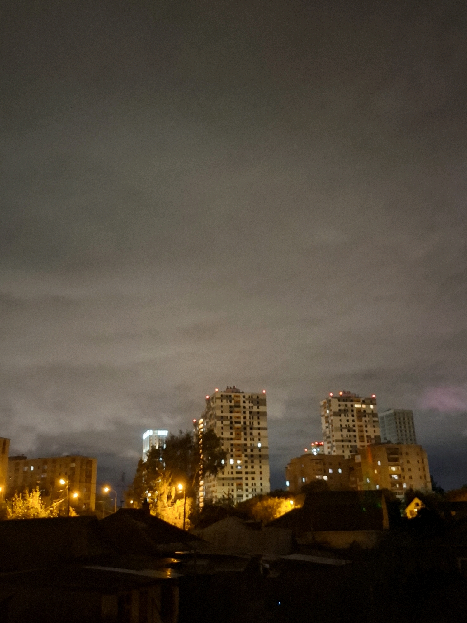 The image depicts a nighttime cityscape, likely in Tyumen, Russia, characterized by a cloudy sky illuminated by artificial lights from buildings and streetlamps. Several multi-story apartment buildings are visible, with lights on in many windows, suggesting residential occupancy. The foreground is dominated by dark, silhouetted rooftops, possibly of lower-rise structures or industrial buildings, with some reflections on wet surfaces indicating recent precipitation. Streetlights cast a warm, yellow glow on the surrounding trees and foliage, which appear dense in some areas. The overall atmosphere is one of an urban environment at night, with a subdued, slightly overcast sky that mutes the intensity of the lights. No people or vehicles are discernible in the frame. The buildings are of modern architecture, typical of urban development. There is a slight reddish hue in the upper right portion of the image, possibly due to distant lighting or atmospheric conditions.