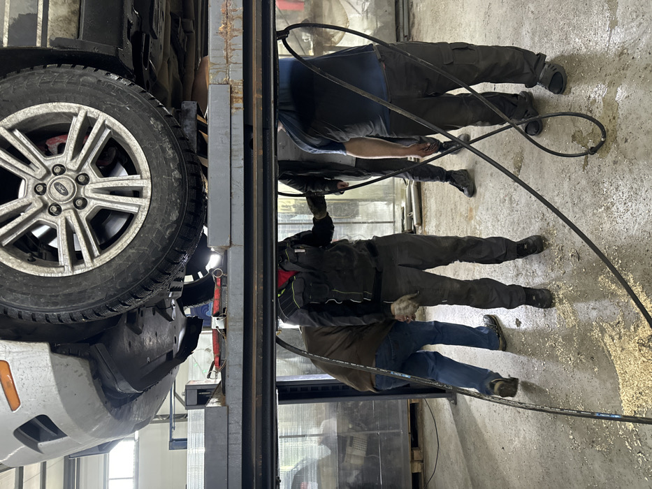 An indoor scene in Zhabkino, Russia depicts three individuals attending to the underside of a silver-colored vehicle, possibly a Ford based on the wheel hubcap, which is elevated on a metal hydraulic lift. The vehicle's front passenger side, including a multi-spoke alloy wheel with a winter tire and a red brake caliper, is prominently visible. Below the lift, a concrete floor is scattered with sawdust or wood chips and shows patches of moisture.
From left to right:
1. A male individual is partially visible, wearing a brown long-sleeved shirt and blue jeans, bending forward, facing away from the viewer. Black athletic shoes are worn.
2. A male individual stands centrally, wearing a dark grey or black work suit with yellow-green reflective accents on the shoulders and arms, dark work gloves, and black work boots. His head is not visible, obscured by the lift structure. He appears to be holding or examining something near his waist.
3. A male individual stands on the right, wearing a dark blue t-shirt, dark cargo pants, and black slides/sandals. His head is also not visible. He holds a black hose or cable in his left hand, which extends to the floor. Another male figure in a dark jacket and pants is partially visible behind this individual.
Multiple black hoses or cables traverse the floor around the individuals, suggesting active work. Red-handled tools are visible on the lift platform beneath the vehicle's front fender. The background includes what appears to be fluorescent lighting and transparent or reflective panels, indicative of a workshop or garage environment.