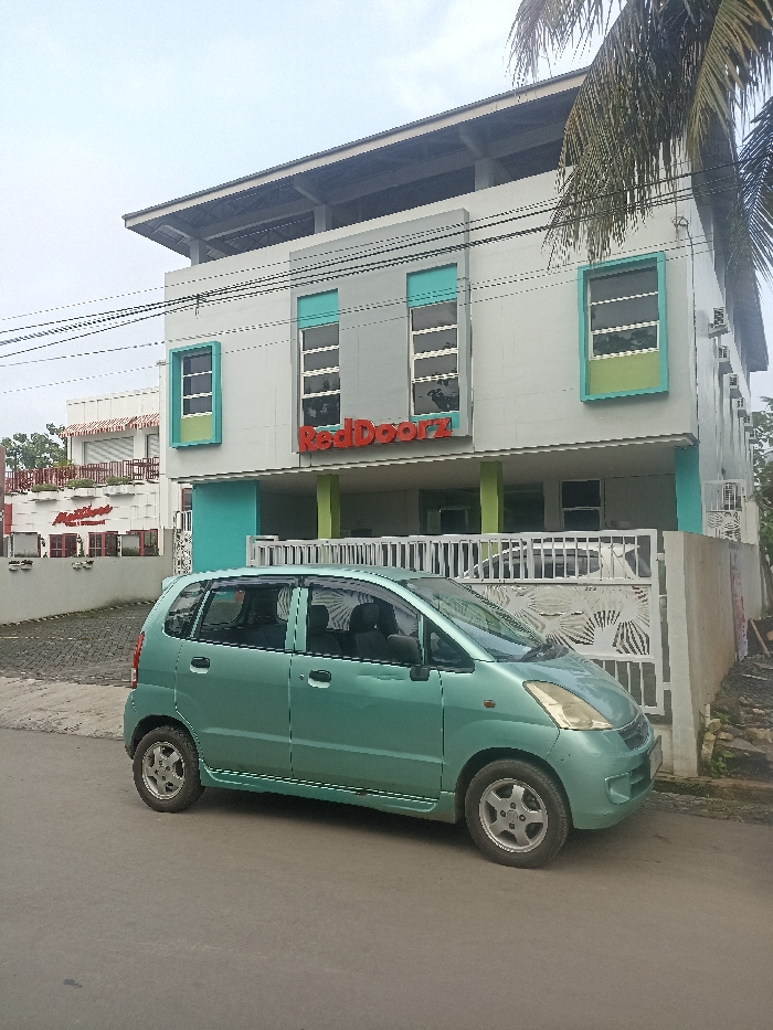 A light green Suzuki Karimun Wagon R is parked in front of a two-story building in Purwokerto, Indonesia. The building is painted white, with teal accents and windows. There is a large, red "RedDoorz" sign on the front of the building. The building appears to be a lodging establishment. The car is facing to the right and there are no people in the image. The ground is black asphalt and the sky is gray with a few white clouds. The image appears to have been taken on a cloudy day. A second building with a red awning is located to the left of the building in the image, and it appears to be a restaurant or cafe. There are many power lines running across the scene, in front of the building. A small, white palm tree is visible to the right of the building. The scene is typical of a street in a small town in Indonesia, with the main point of interest being the car and the building. A metal gate is positioned between the car and the building.