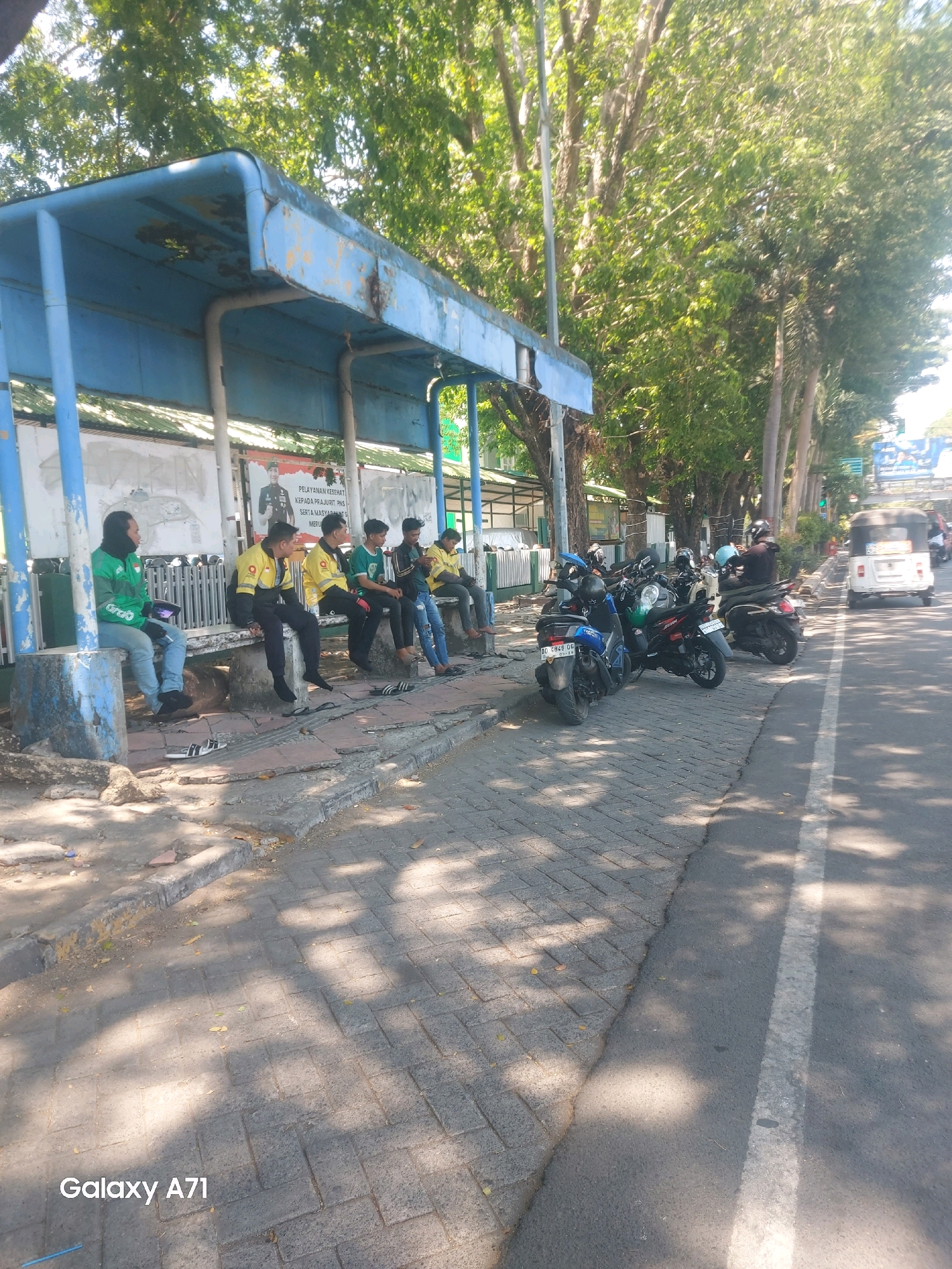 The image depicts a street scene in Makassar, Indonesia, likely near a bus stop or public waiting area. Several individuals, mostly young men, are seated on a bench beneath a blue, weathered bus stop shelter. Some are wearing brightly colored jackets, suggesting they might be part of a delivery service or sports team. Motorbikes are parked along the sidewalk, indicating a common mode of transportation in the area. The street itself is paved with interlocking bricks on the sidewalk and asphalt on the road, with a white line marking the lane. Lush trees line the street, casting dappled shadows on the ground, suggesting a warm, sunny day. In the background, a white, three-wheeled vehicle, possibly a bemo or a rickshaw, is visible on the road, along with other traffic. Posters are displayed on the wall behind the bus stop. The overall impression is of a typical urban public space in Makassar.