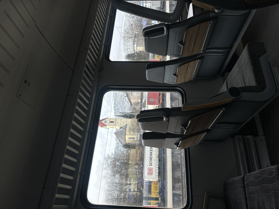 The image depicts the interior of a passenger train car, looking outward through a window at a railway environment adjacent to the city of Kufstein, Austria.
Inside the train, multiple rows of unoccupied grey passenger seats with wood-grain accent panels are visible. The ceiling features light-colored panels with linear ventilation grilles and overhead fixtures. A heater unit is positioned on the floor beneath the window.
Outside the window, multiple parallel railway tracks are present. Several freight cars are parked on these tracks, including a white "DB SCHENKER" branded car, a yellow car, and a red car further down the line. Beyond the railway, the prominent Stadtpfarrkirche St. Vitus, identifiable by its yellow facade and distinctive red-capped steeple, stands on an elevated section of the city. Additional buildings, including multi-story structures, are visible around the church. In the background, snow-dusted mountains or hills rise under an overcast sky, with bare deciduous trees and some evergreens dotting the landscape. No individuals are visible inside or outside the train.