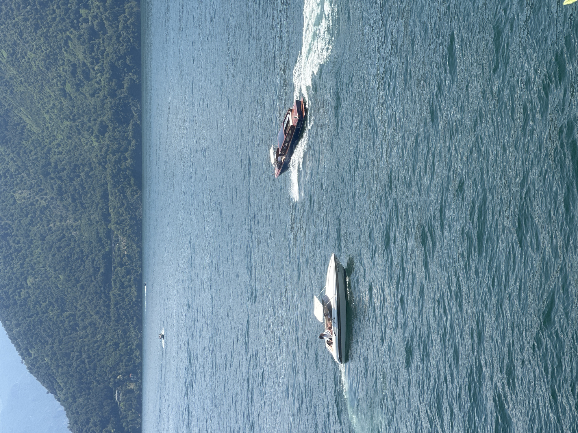 This is a high-angle, full shot of a lake in Tremezzina, Italy, with a heavily wooded mountain range in the background. Two boats are visible on the water. In the foreground left, a white motorboat with a canopy top is moving to the left, leaving a wake. Three individuals are seated in the boat. In the middle ground right, a dark blue motorboat with a red stripe and a white canopy is moving to the right, also leaving a wake. The water is choppy with light reflecting off the surface. The mountain range is covered in dense green trees and appears steep. The sky is clear and bright.