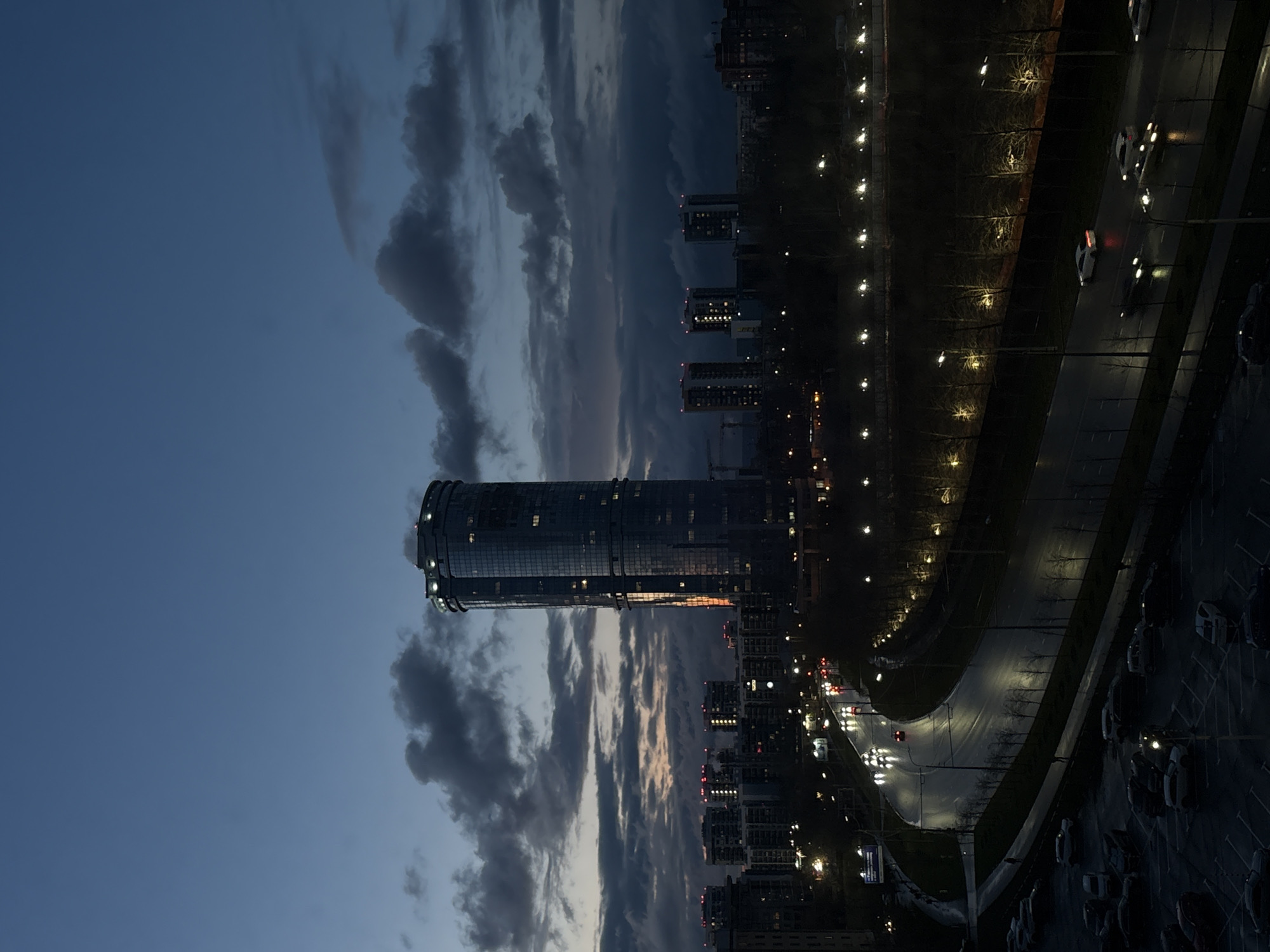This image presents an urban landscape of Kazan, Russia, at dusk or early night, viewed from a high vantage point. The sky occupies the upper left portion, a deep twilight blue adorned with scattered, dark clouds. A faint, warm glow from the fading sunset can be seen along the horizon line in the middle distance.
A prominent, modern, cylindrical glass skyscraper dominates the left side of the frame, its structure subtly illuminated by interior lights and a few decorative exterior lights at its crown and base. Below and to its right, several other multi-story buildings, likely residential, dot the cityscape, some showing illuminated windows. Faint outlines of construction cranes are visible near the base of the main skyscraper.
To the right, a wide, multi-lane road curves through the scene, its surface appearing reflective, possibly due to recent rain or the strong reflection of artificial lights. Cars with illuminated headlights and taillights are visible on the road, indicating active traffic. Numerous streetlights brightly illuminate the road and a strip of bare trees lining its edge, suggesting late autumn or winter. Further to the bottom right, a parking lot is densely packed with parked vehicles.
The scene conveys a quiet, contemplative mood as the city transitions from day to night. No people are clearly visible, and there is no legible text within the image.