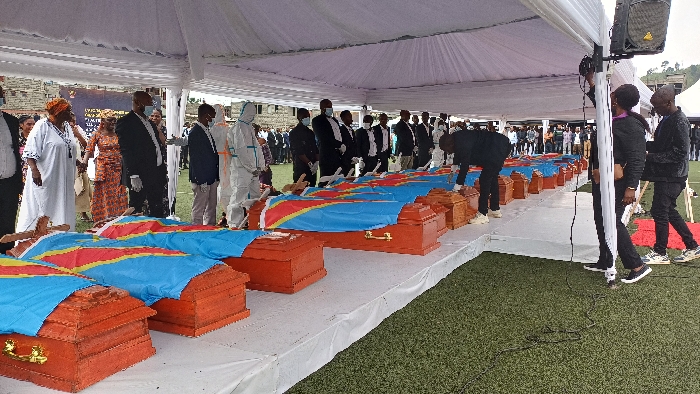 Approximately eighteen reddish-brown wooden caskets, each draped with the national flag of the Democratic Republic of the Congo, are arranged in a single long row on a white-covered platform. This scene takes place outdoors on a green artificial turf field in Goma, Democratic Republic of the Congo, beneath a large white canopy tent. A group of individuals stands alongside the caskets, observing solemnly. This group includes persons in formal dark suits, traditional African attire, and at least two individuals wearing full white protective personal equipment (PPE) hazmat suits with face masks. One man in a black suit and white sneakers is bending over a casket on the right side of the formation. In the background, beyond the covered area, a larger gathering of people is visible. Hilly terrain and buildings can be seen further in the distance. Audio equipment, including a speaker, is mounted on a tent pole on the right.