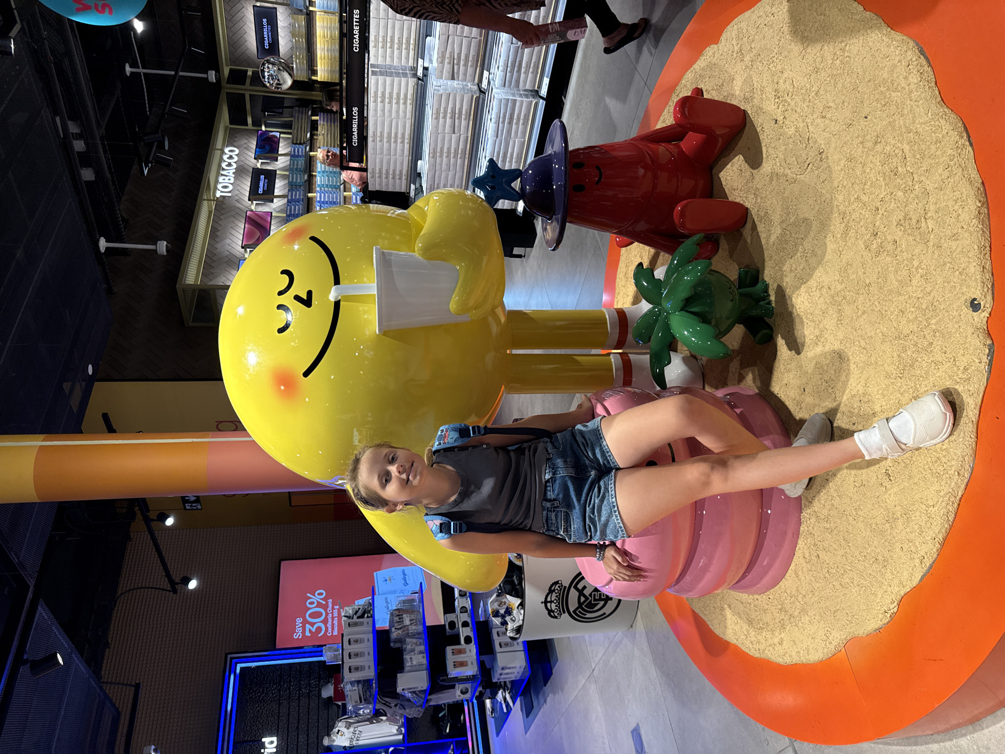 A young girl with brown hair pulled back, wearing a gray tank top and denim shorts, sits on a large pink, stacked object with a Real Madrid logo on a white cylindrical object behind her. She is positioned in front of a giant yellow cartoon character that holds a white cup. To the right of the yellow character, a small red cartoon figure wearing a blue hat with a star sits on a sandy surface surrounded by an orange circular border. In the background, shelves are stocked with various products, and a "TOBACCO" sign is visible. The overall setting appears to be an indoor retail space, possibly a souvenir or convenience store.