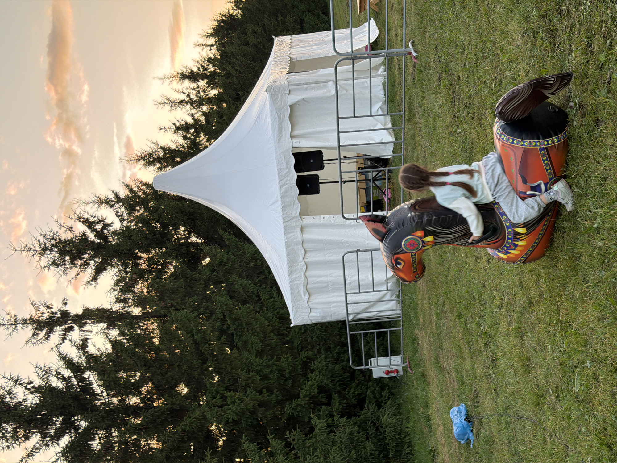 A young girl with braided pigtails sits on an inflatable horse toy in a grassy field. She is facing away from the camera. Behind her, a large white tent with a peaked roof is set up, with two black speakers on stands in front of it. Trees line the background of the scene. The sky is a soft orange and pink, indicating sunset or sunrise. A blue plastic bag lies on the grass to the left of the inflatable horse.