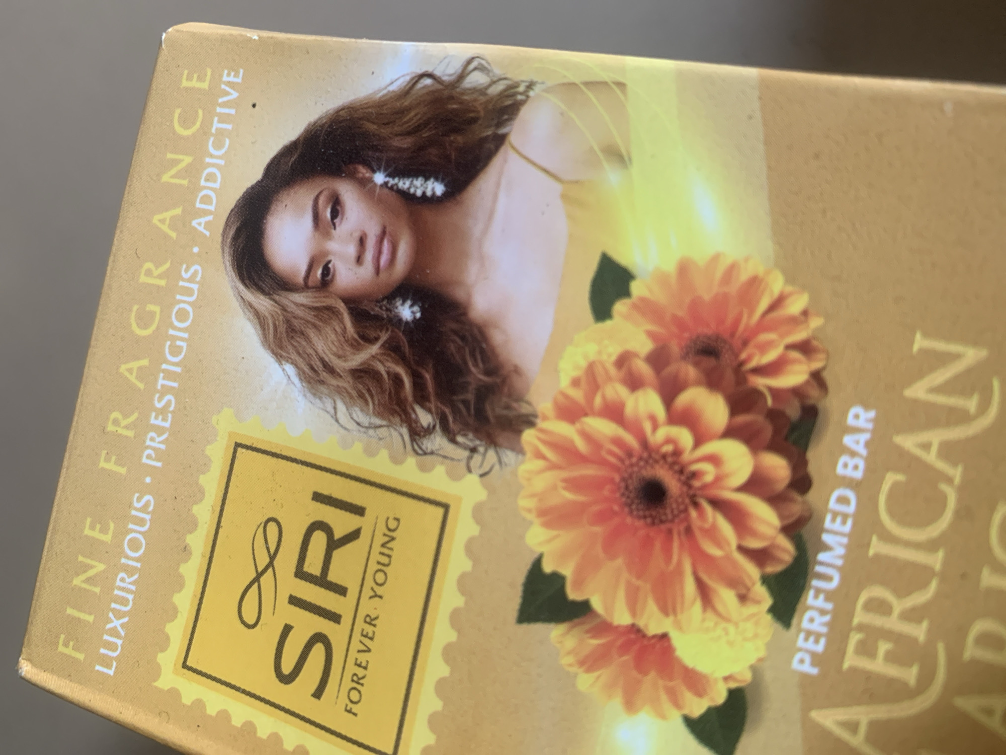 The image is a close-up of the packaging for a product labeled "SIRI FOREVER YOUNG PERFUMED BAR AFRICAN FABRIC." The packaging is predominantly yellow and features text such as "FINE FRAGRANCE," "LUXURIOUS," "PRESTIGIOUS," and "ADDICTIVE." A portion of a woman's face and shoulders is visible, with wavy brown hair and dangling earrings. In the foreground, there are several orange and yellow flowers. The product is likely a soap bar, given the "PERFUMED BAR" text. The image is taken from a slightly elevated angle, focusing on the product packaging. There is no identifiable information or context within the image to associate it with Jauro Yinu, Nigeria.