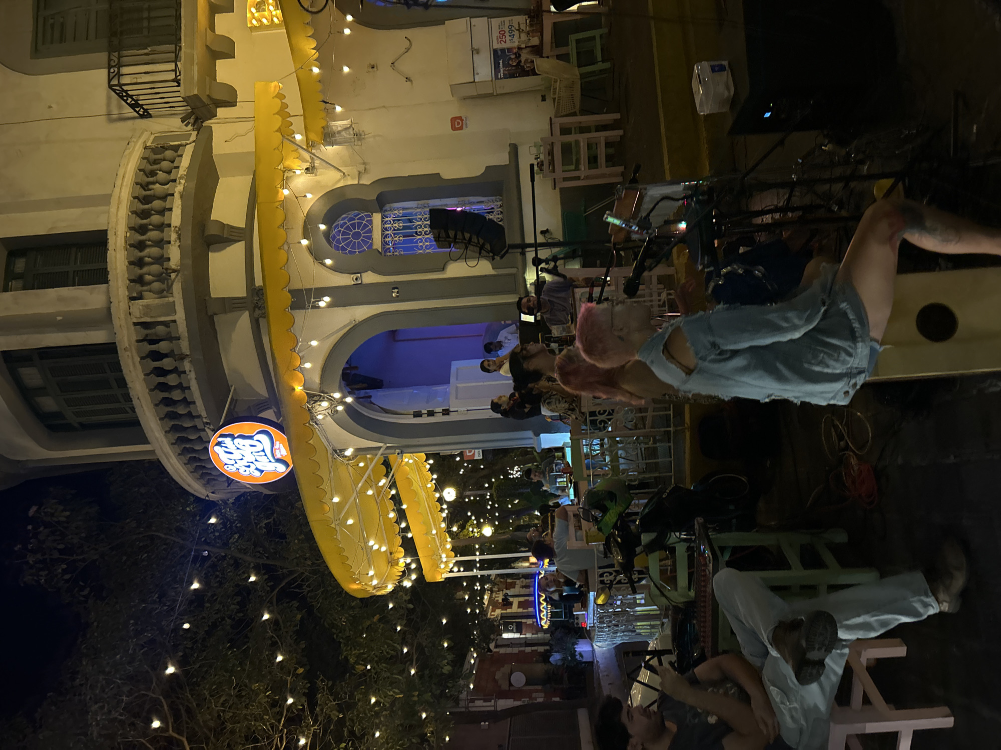 The image shows a nighttime scene at a restaurant or bar in Mazatlán, Mexico. A yellow awning with lights covers an outdoor seating area. People are seated at tables, and others are standing and interacting. The restaurant is decorated with string lights in the trees. The building has architectural details, including arched windows and a rounded tower. A sign with the restaurant's name is visible. People are casually dressed. Some patrons are seated in chairs. It is a lively social atmosphere.