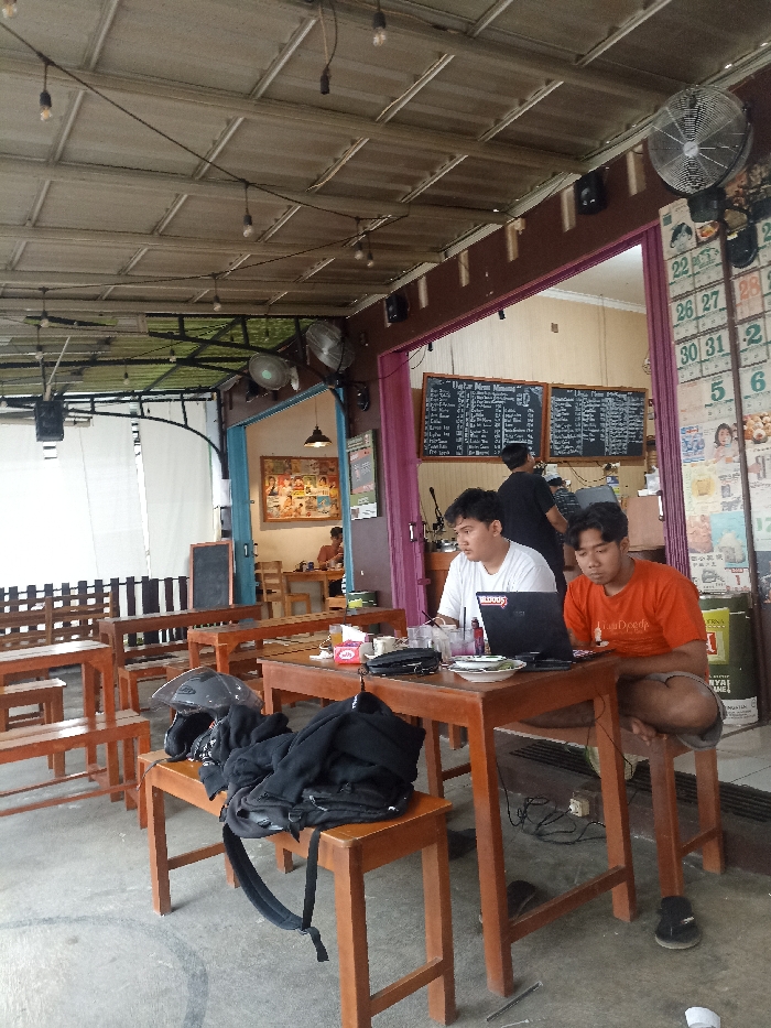 The scene is an outdoor seating area of a restaurant in Purwokerto, Indonesia, located at the coordinates -7.425 latitude and 109.228 longitude. There are three men sitting at wooden tables. One man is using a laptop computer. A second man is seated at a table in front of him, holding a coffee cup and looking at the laptop. There is a third man sitting at another table, but he is obscured from view. The seating area is covered by a corrugated metal roof and a clear plastic awning. There are string lights overhead, a ceiling fan, and a few potted plants visible in the background. The wooden tables are surrounded by wooden chairs and a few wooden benches. A backpack is sitting on one of the benches. There are also some items on the tables, including a coffee cup, a plate, and a laptop computer.