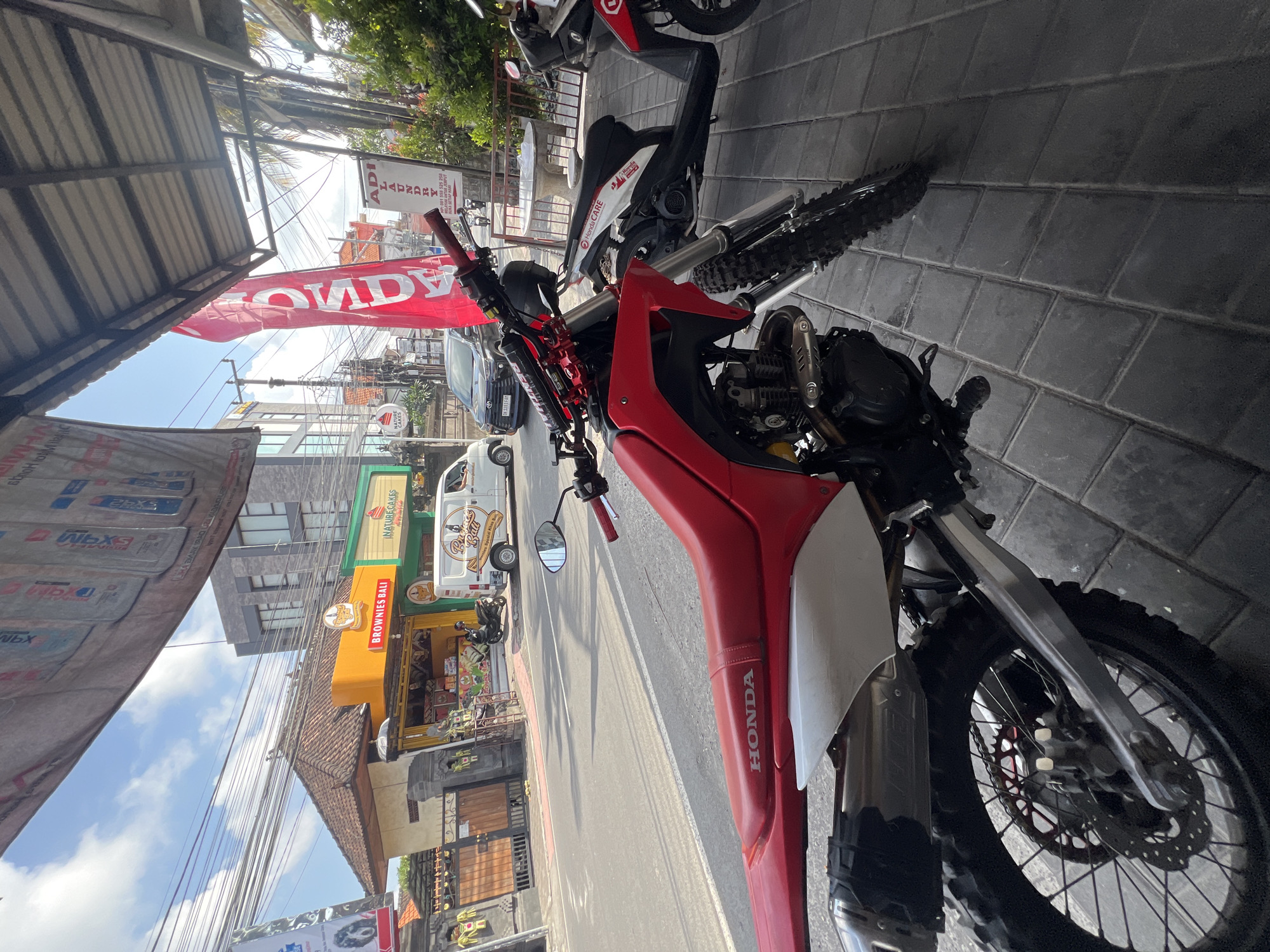 A red and white dirt bike is parked on a paved sidewalk in front of a row of shops in Denpasar, Indonesia. The bike has a silver exhaust pipe and black tires. In the background, a white van with "Brownies Bali" written on the side is parked on the street. The street is lined with shops selling food and drinks. A red flag with "HONDA" written in white letters is also visible. The sky is blue with some clouds.