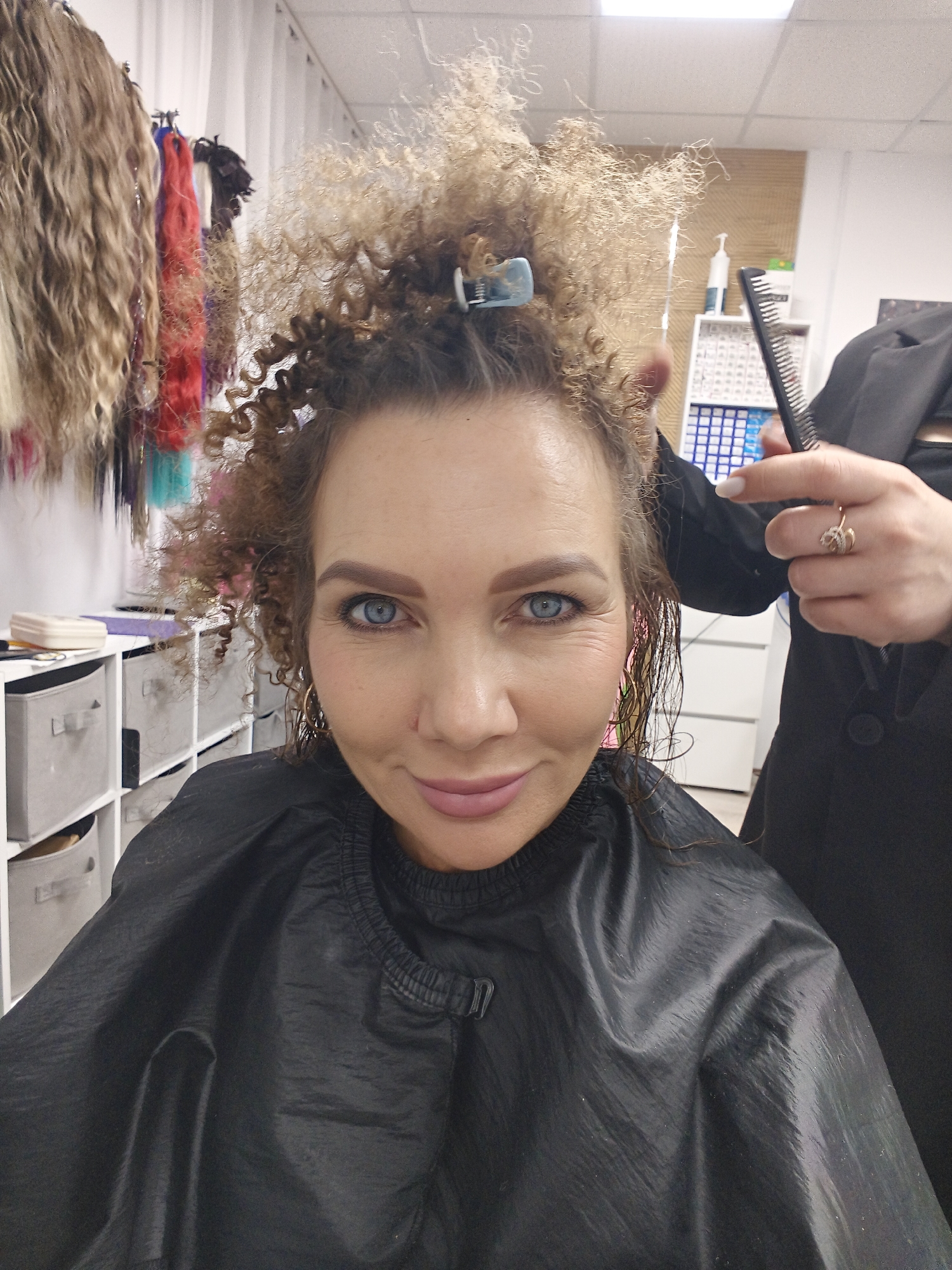 A medium shot captures a woman smiling directly at the camera within what appears to be a hair salon. She has striking blue eyes and light brown, very curly hair, much of which is sectioned and clipped on top, while other strands appear wet. She wears a black, shiny salon cape that covers her shoulders and chest, secured around her neck. A small gold hoop earring is visible on her left ear.
To the right, the hand of a stylist is partially visible, holding a black comb near the woman's head, suggesting a hair treatment or styling is in progress. The stylist's hand has white nail polish and wears a gold ring with an intricate design on the ring finger.
The background reveals a professional salon setting. On the far left, various colors of hair extensions, including shades of blonde, brown, red, purple, black, and turquoise, hang on a rack. Below these, a white shelving unit holds several gray fabric storage bins. To the right, behind the stylist's hand, another white shelving unit is filled with numerous small compartments, likely containing hair products or dyes, with a pump bottle sitting on top. The wall behind this unit has a textured, woven pattern, possibly wicker. The ceiling consists of white panels with integrated fluorescent lighting, indicating an indoor environment. The woman's cheerful smile conveys a positive mood during the session.