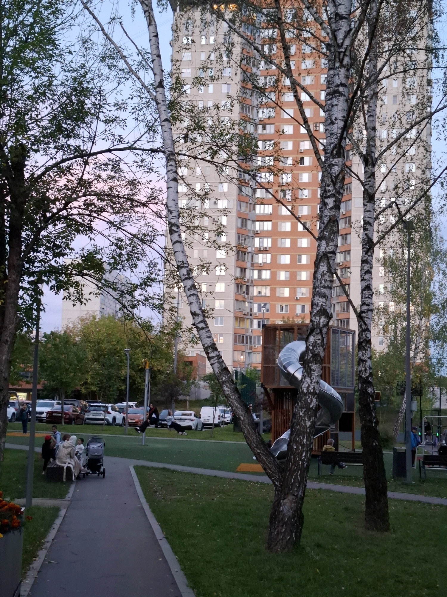 In an urban setting, a large apartment building with many windows dominates the background. In the foreground, trees with white and black bark are prominent. A playground with a silver slide is visible between the trees. A paved path winds through a grassy area where several people are gathered, including adults and children, some sitting on a bench and one with a stroller. Cars are parked in the background. The sky displays a gradient of pale blue to pink, suggesting dusk.